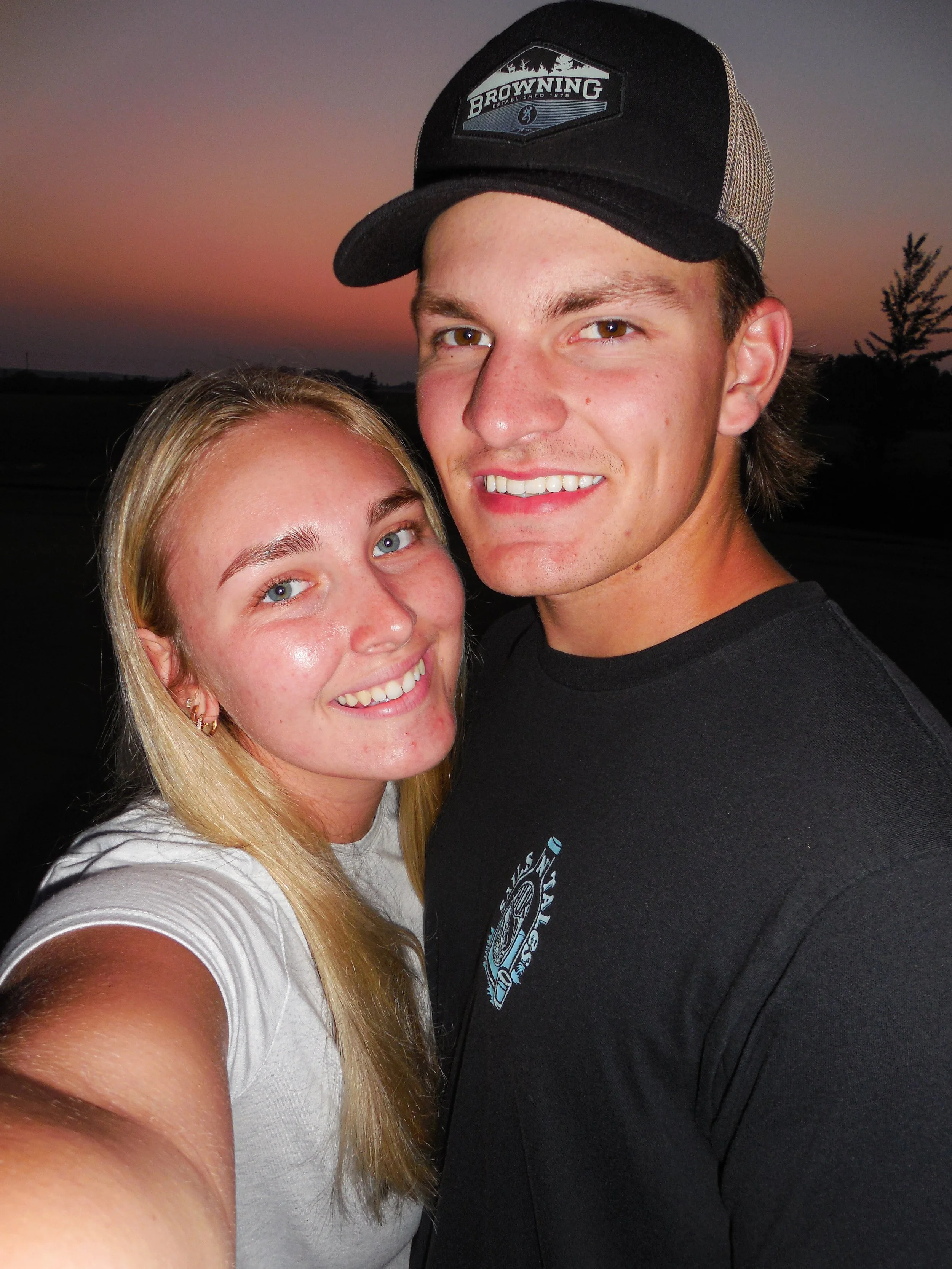 Close-up selfie of a young woman and a young man smiling outdoors at sunset. The woman has long blonde hair and blue eyes, and is wearing a white shirt. The man has short brown hair, hazel eyes, and is wearing a black cap and black T-shirt.