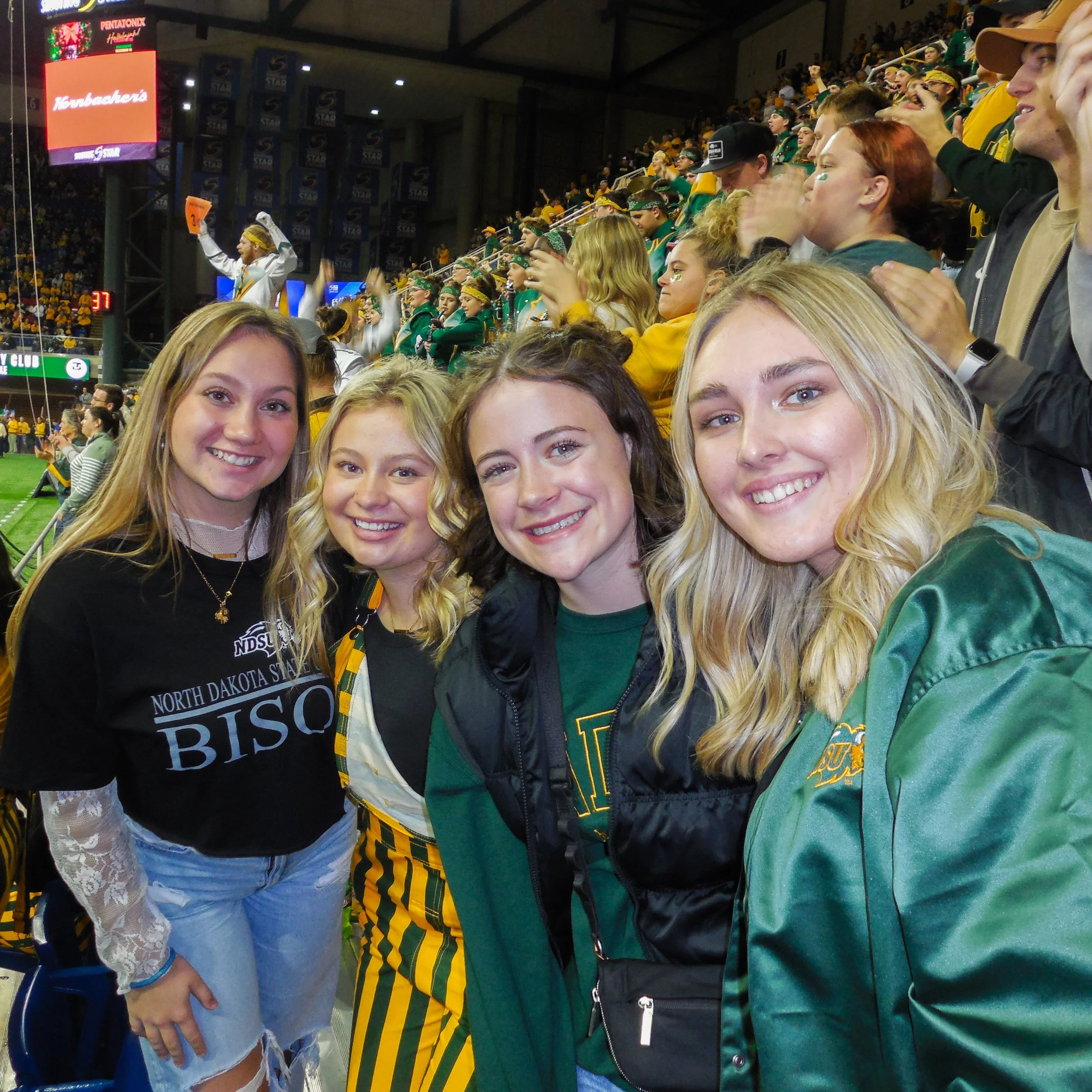 Group of young people at a hockey game, some wearing green and yellow team colors, waving and clapping, with a large crowd in the background.