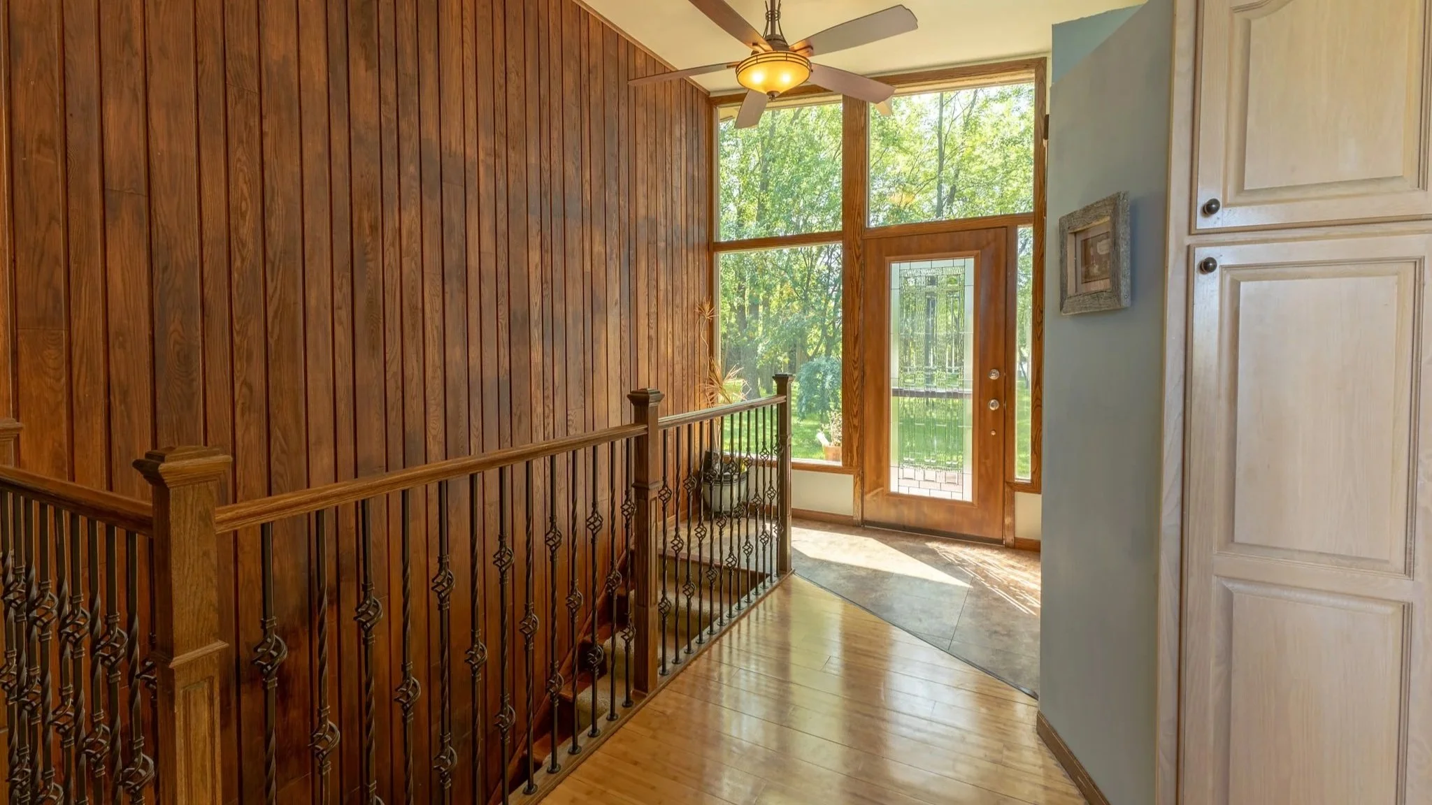 Entryway with large windows, wooden walls, ceiling fan, and front door with glass panel, leading outside to trees.