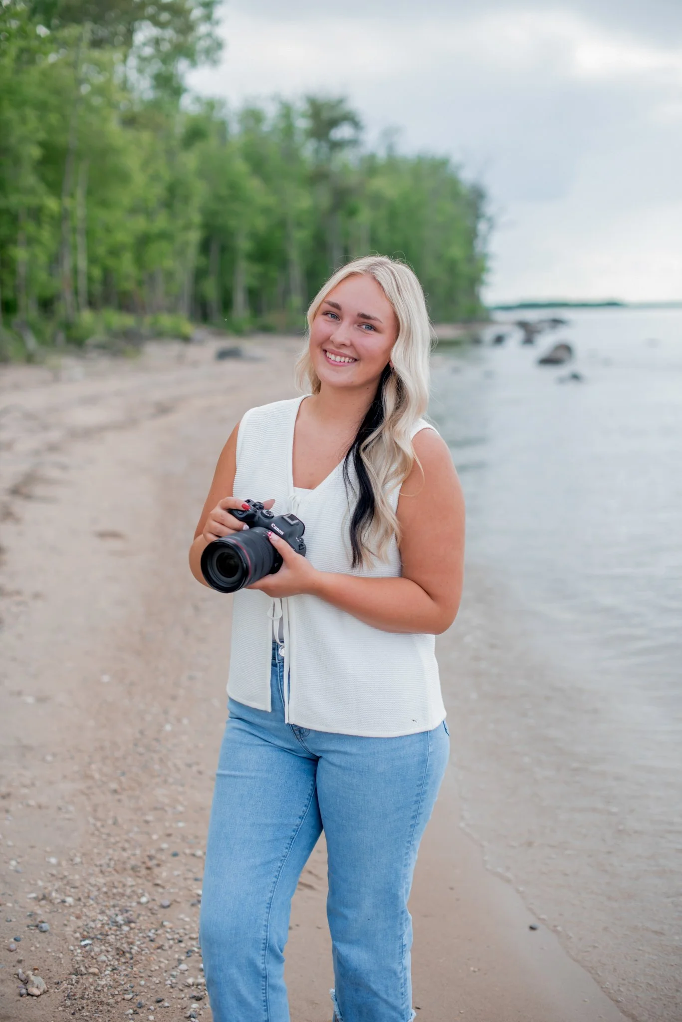 A young woman with long, wavy blonde hair and a big smile holding a Canon camera on a beach with trees in the background.