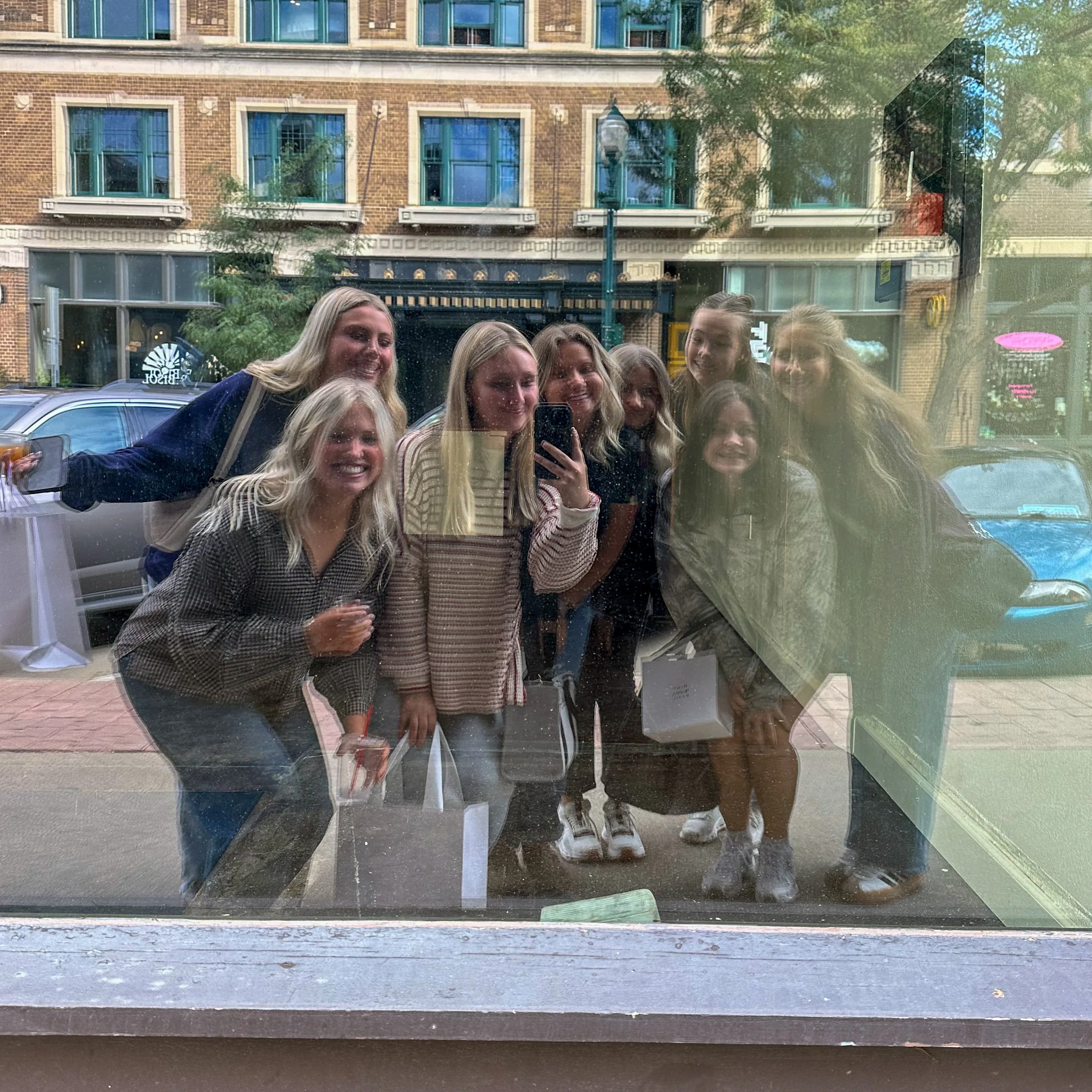 Reflected image of a group of smiling people, possibly friends, taking a photo outside on a street, with urban buildings and trees in the background, visible through a glass window.