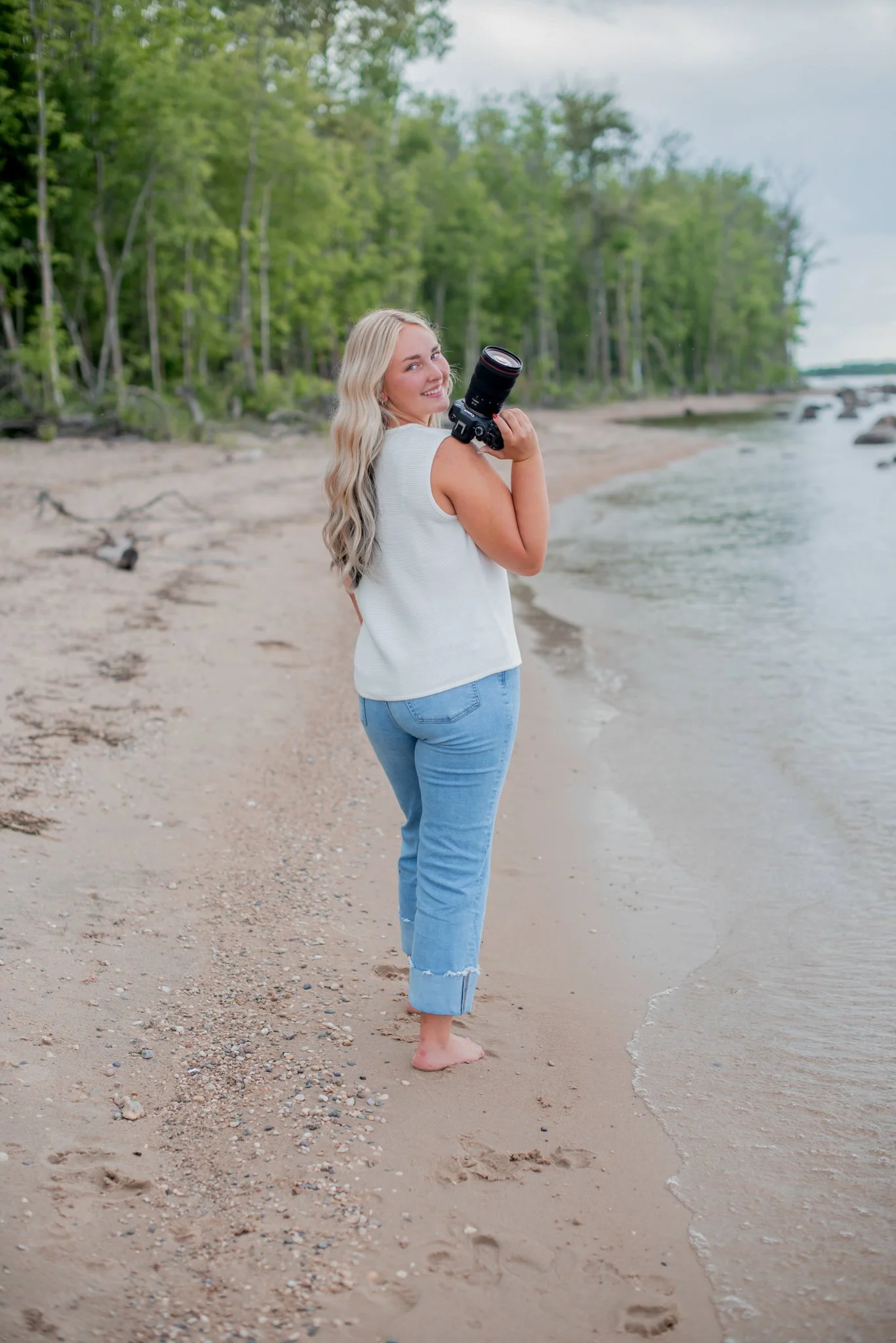 A woman with long blonde hair smiling while holding a camera on her shoulder, standing on a sandy beach near the water with green trees in the background.