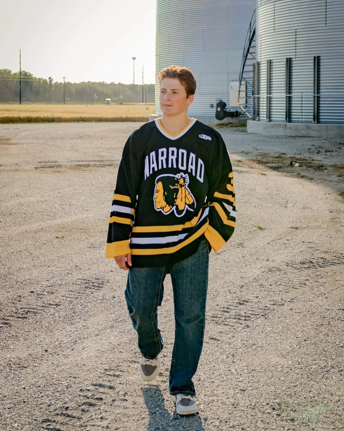A young man wearing a black Marroad hockey jersey and jeans, walking outside on a gravel lot near large metallic storage tanks, with a rural landscape in the background during sunset.