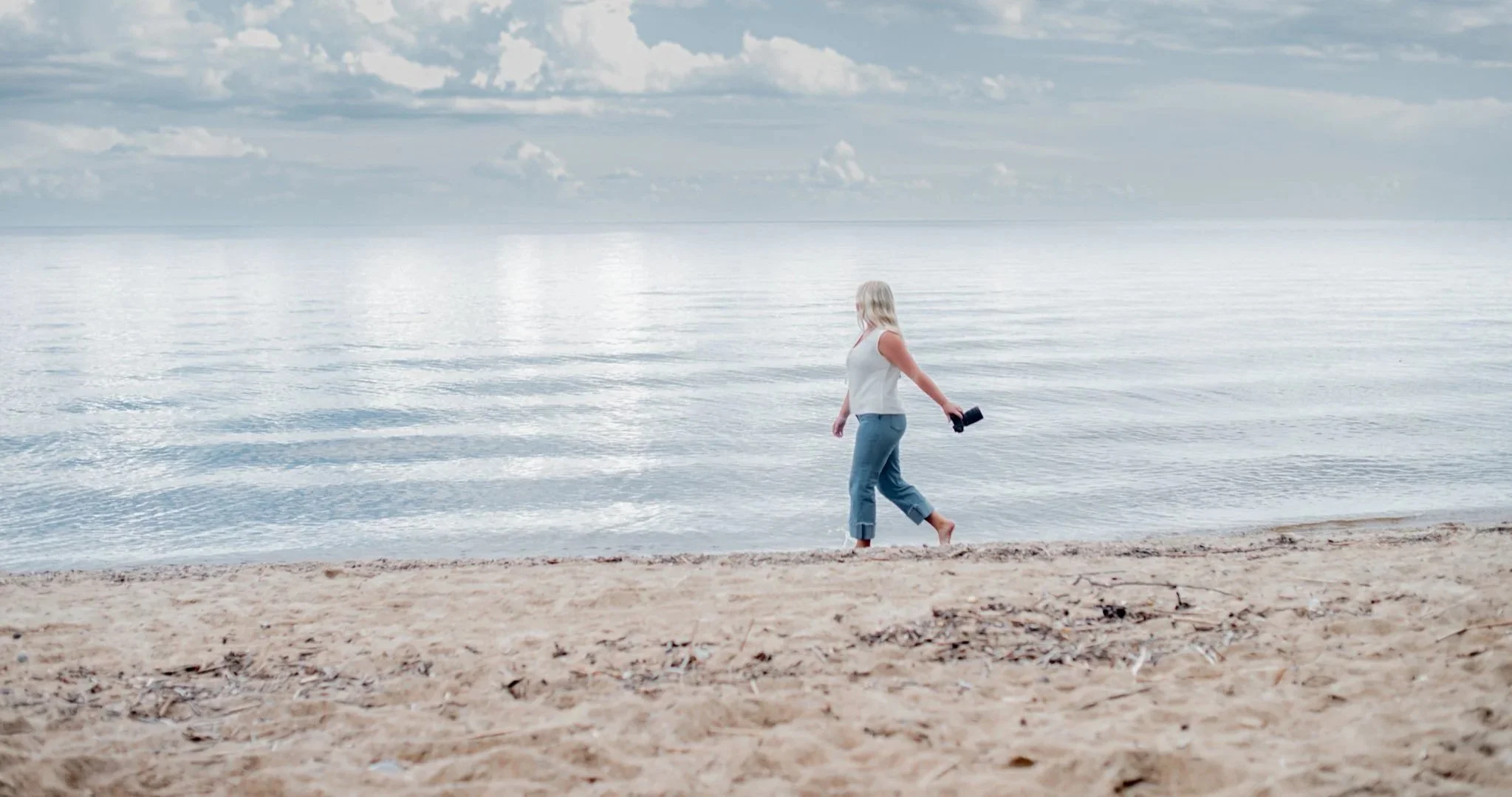 A woman in a white top and jeans walking barefoot on a sandy beach with a camera in her hand, near calm water under a cloudy sky.