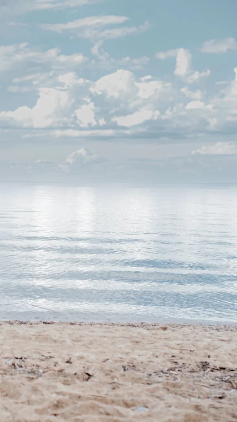 Calm beach scene with light blue sky, scattered clouds, gentle water, and sandy shoreline.