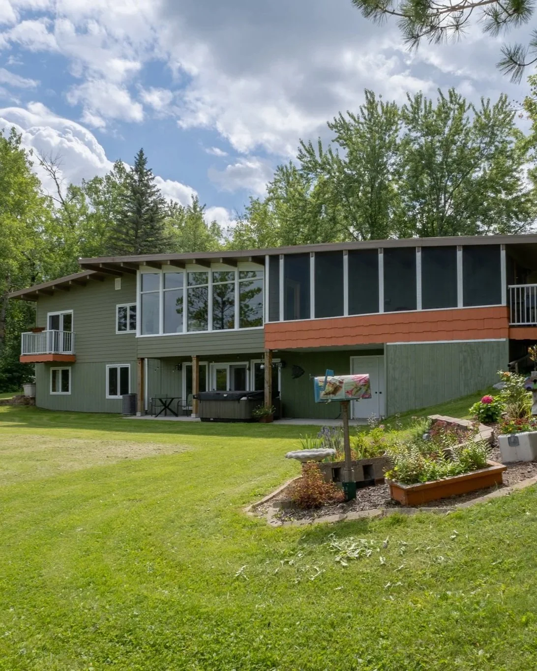A two-story house with green siding, large windows, and a small balcony, surrounded by a lush green yard with garden beds and trees in the background.