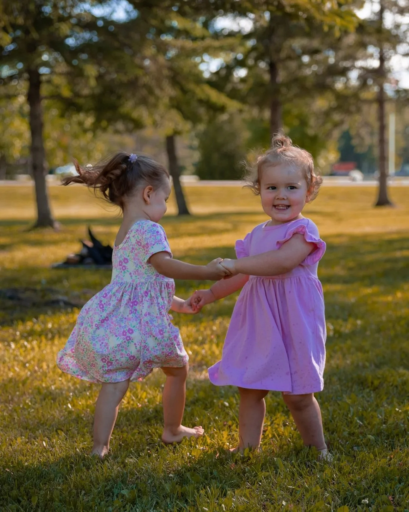 Two young girls in dresses, holding hands and dancing in a park with trees in the background during sunset
