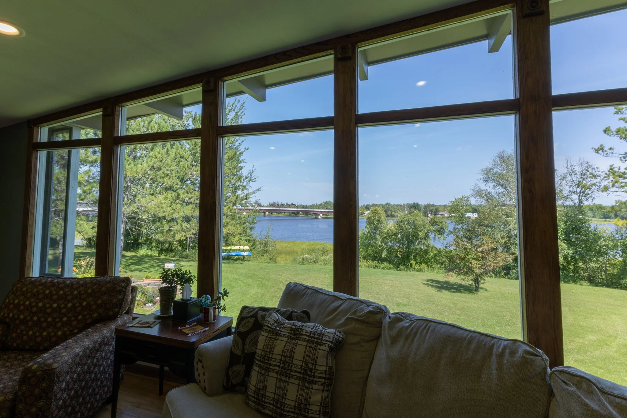 Interior view of a living room with large wall of windows overlooking a lush green yard and a body of water in the distance, under a clear blue sky.