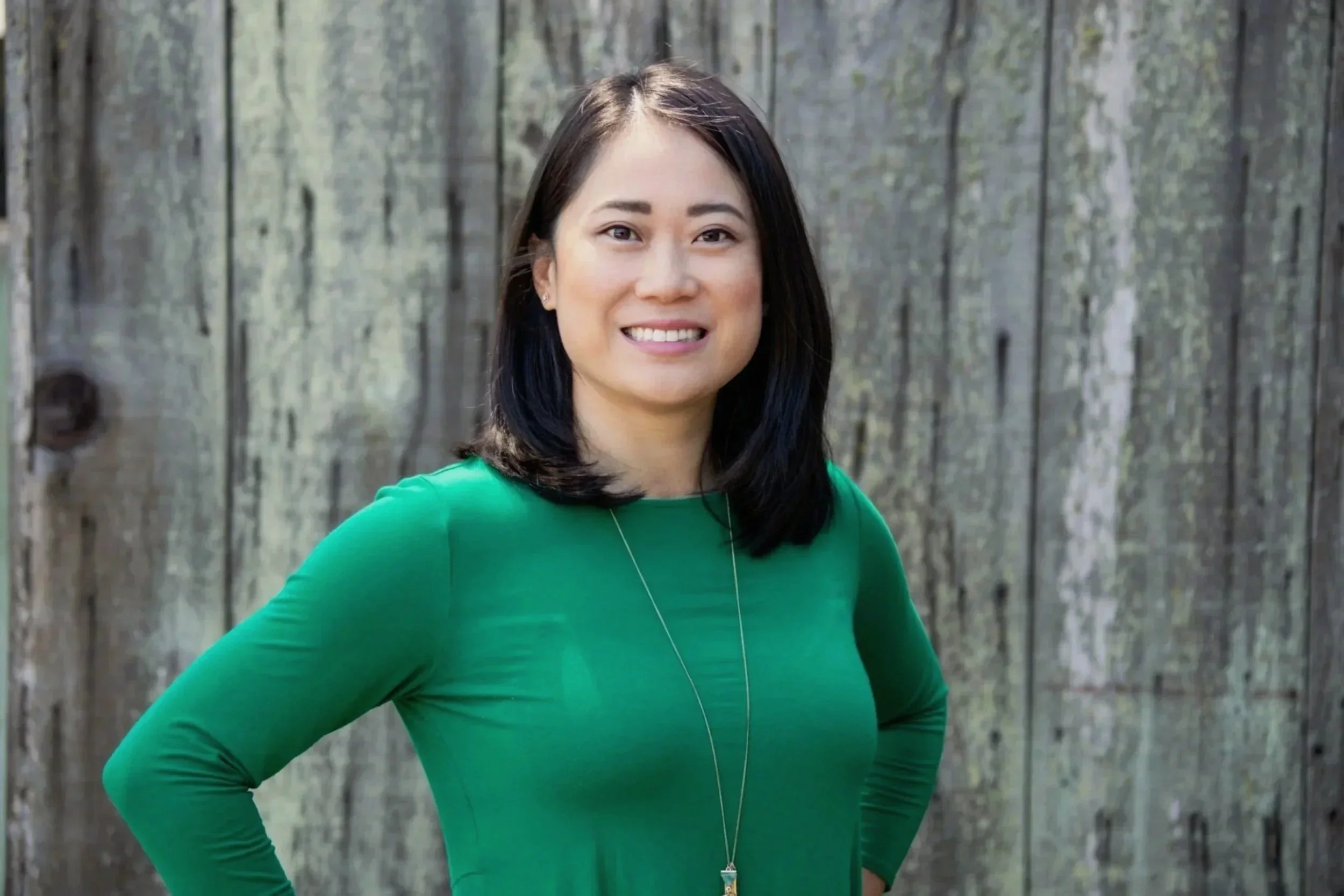 A woman with black hair, wearing a green long-sleeve shirt, standing outdoors in front of a wooden fence, smiling at the camera.