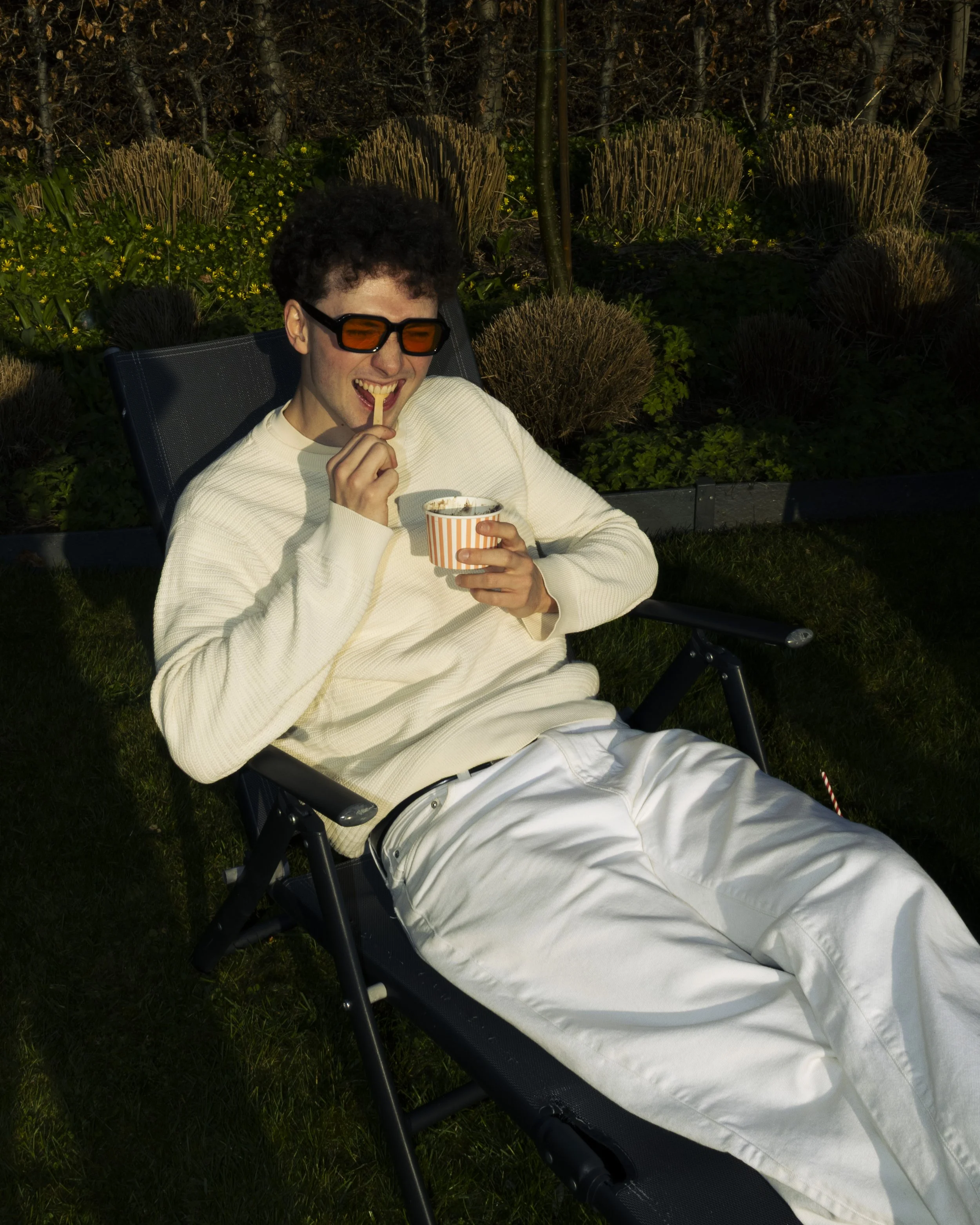 A young man with curly hair and sunglasses sitting in a lounge chair outdoors, eating ice cream from a striped cup. He is wearing a cream-colored sweater and white pants, with a background of yellow flowers, bushes, and a wooded area.