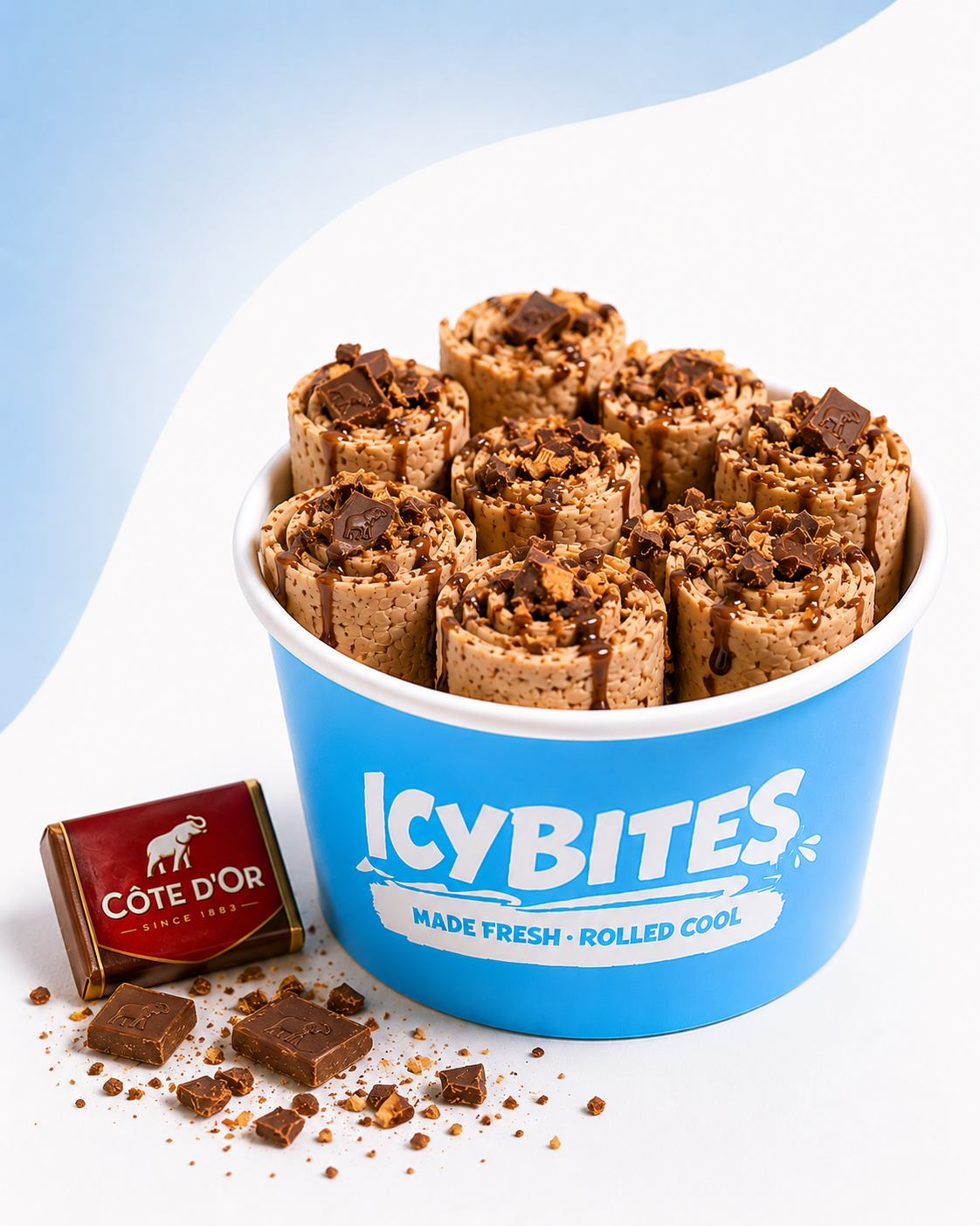 A blue bucket labeled "ICYBITES Made Fresh Rolled Cool" filled with rolled ice cream treats topped with chocolate and chocolate drizzle. A small box of Côte D'Or chocolate and some broken pieces of chocolate are placed beside the bucket on a white surface.