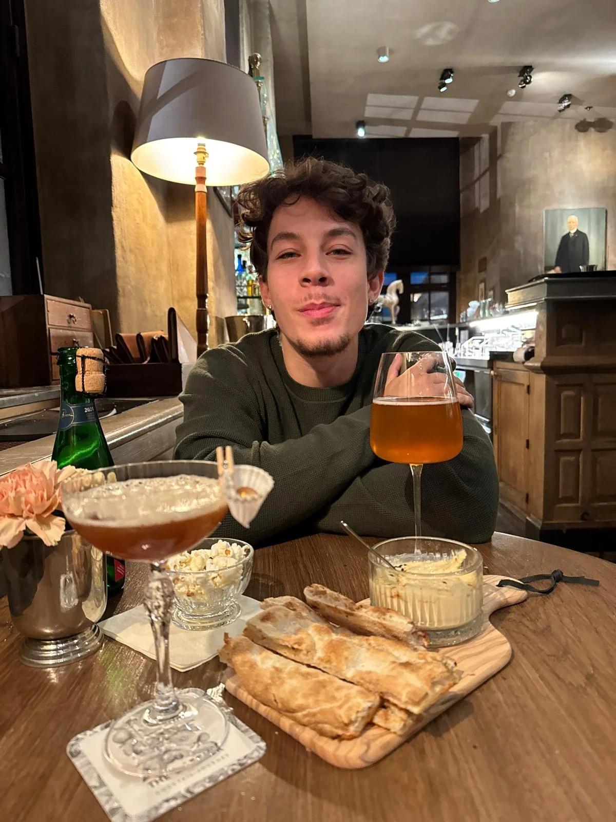 A young man with curly hair and a goatee sitting at a restaurant table with drinks and food, including a cocktail glass with an orange-colored drink, a glass with popcorn, and a wooden tray with flatbread, with warm lighting and wooden decor.