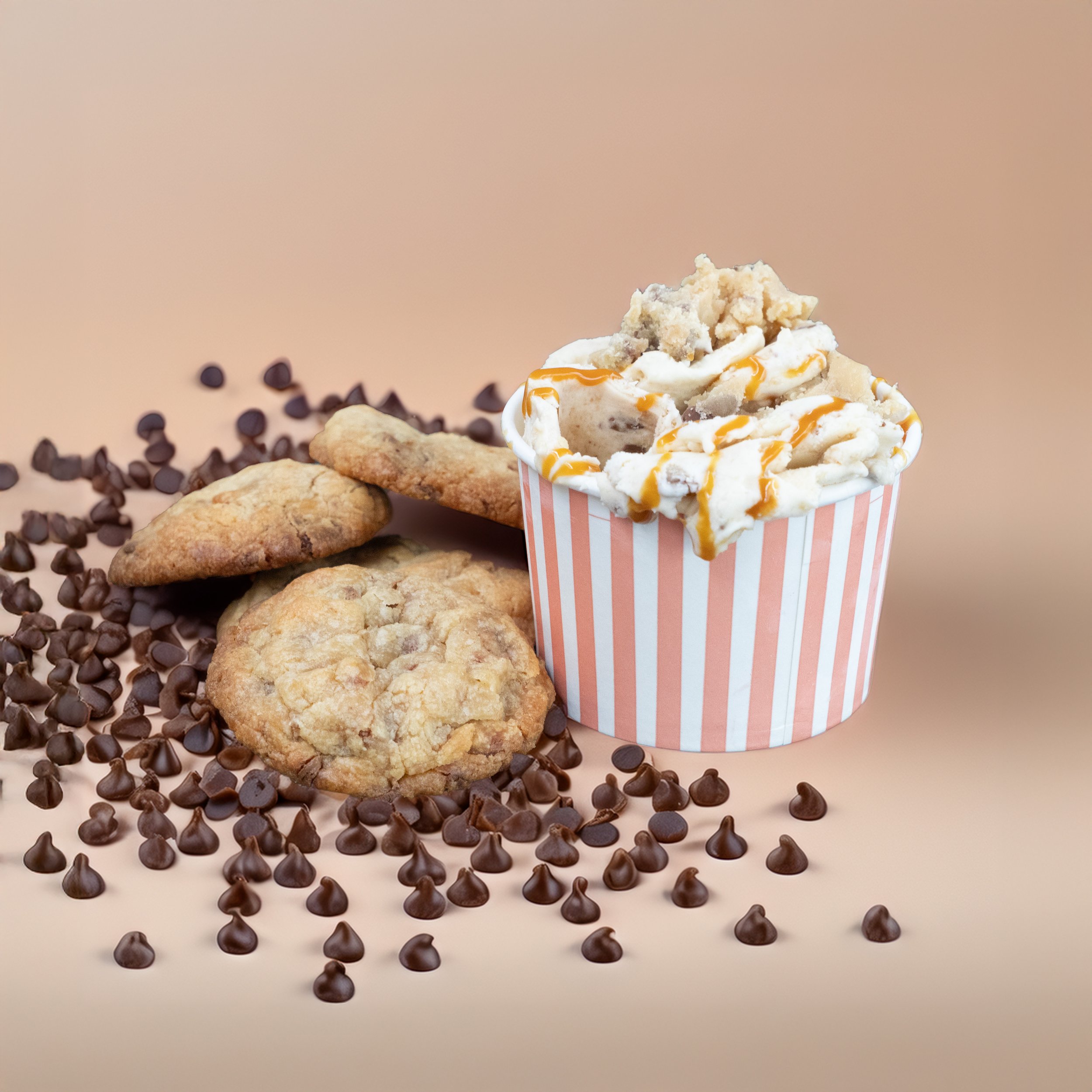 A group of chocolate chip cookies and a cup of ice cream topped with caramel, whipped cream, and cookie crumbs, surrounded by scattered chocolate chips on a beige background.