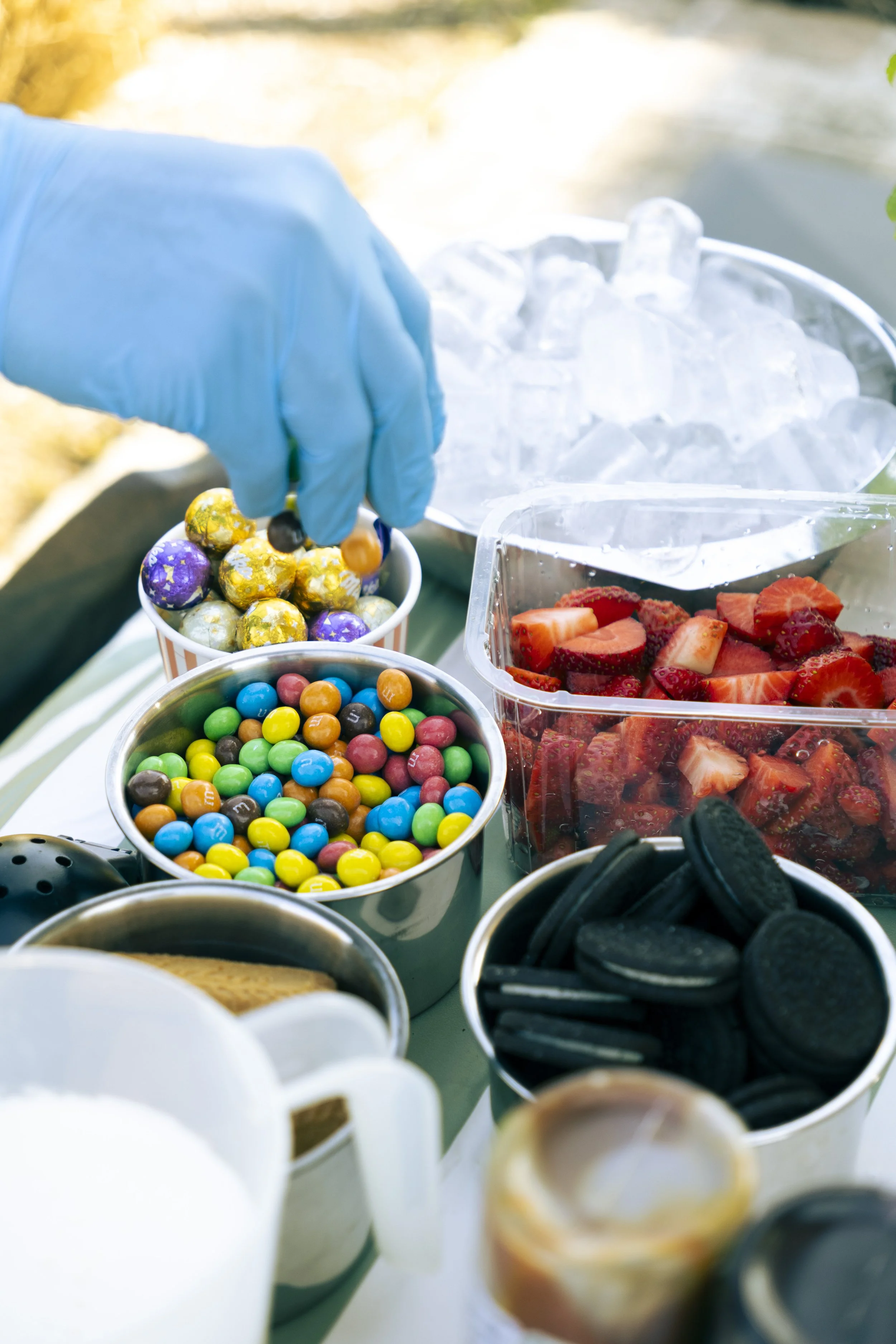 Various bowls of colorful candies and strawberries on a table, with a person wearing a blue glove reaching into one of the bowls.