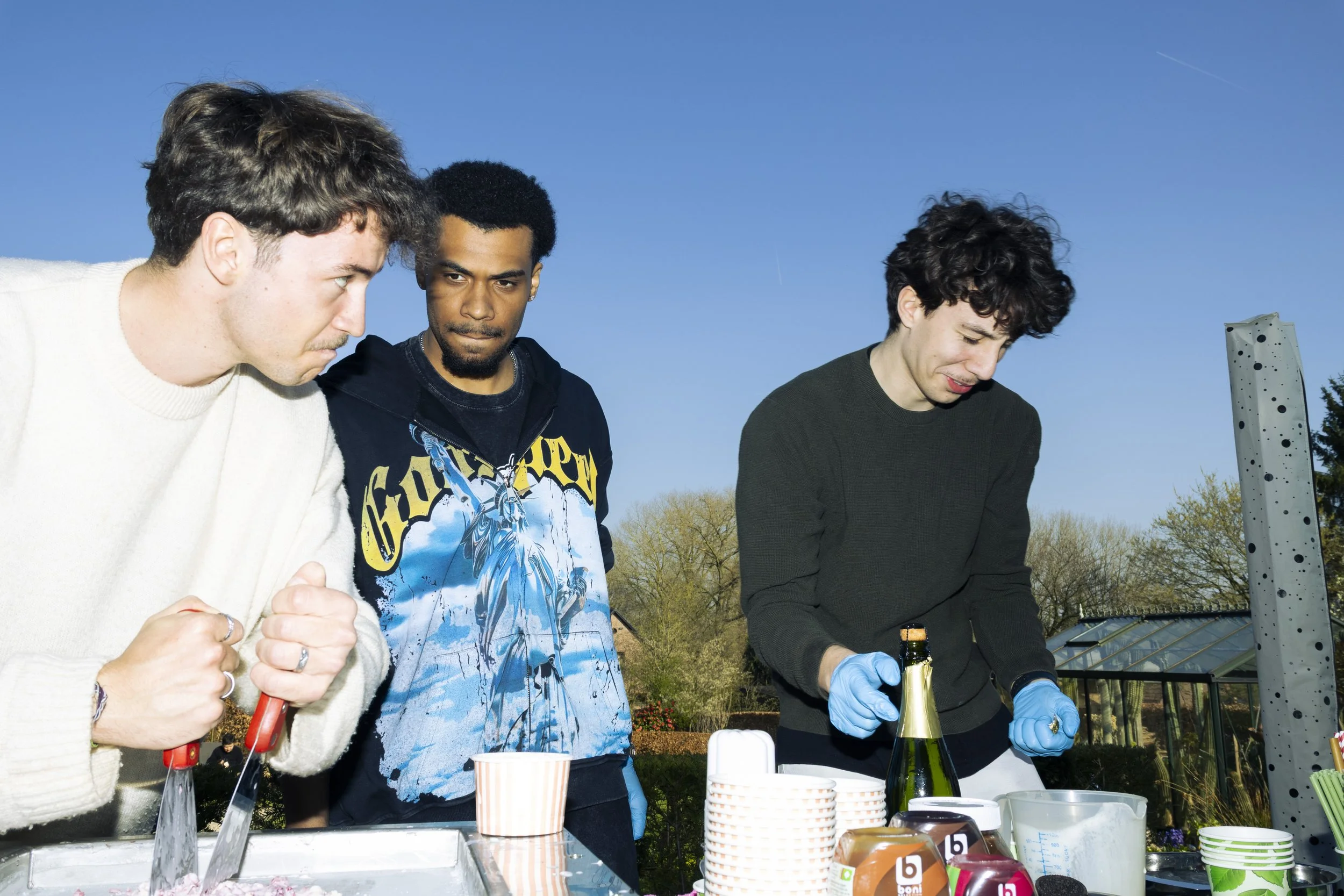 Three men are outdoors, preparing food or drinks at a table with cups and bottles, under a clear blue sky.
