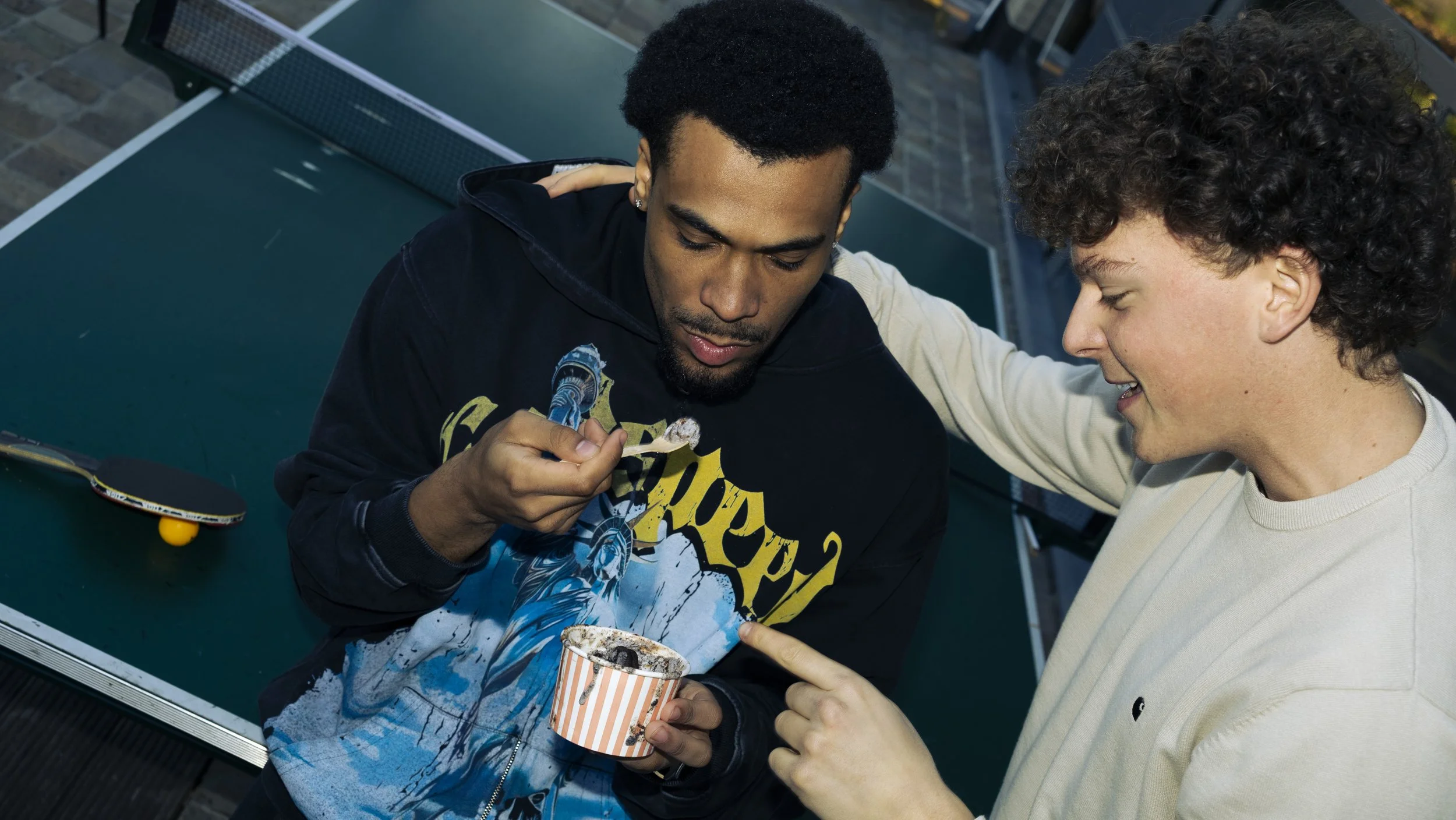 Two young men sharing a moment of enjoyment with one holding a striped cup of ice cream and the other using a spoon to eat from it, sitting outdoors at a table.