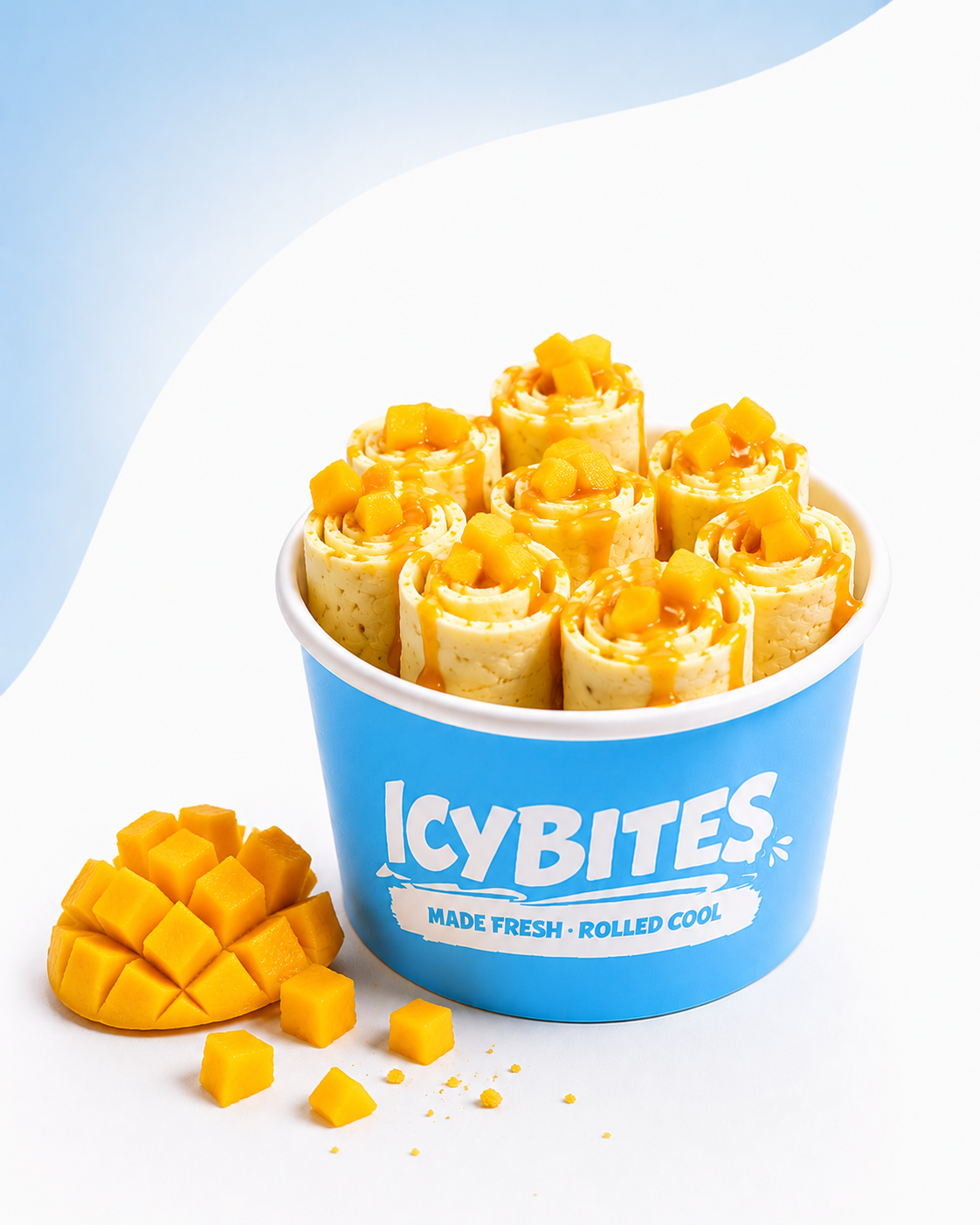 A blue container labeled 'ICY BITES' filled with rolled ice cream topped with mango chunks and mango syrup, with additional mango pieces beside it.
