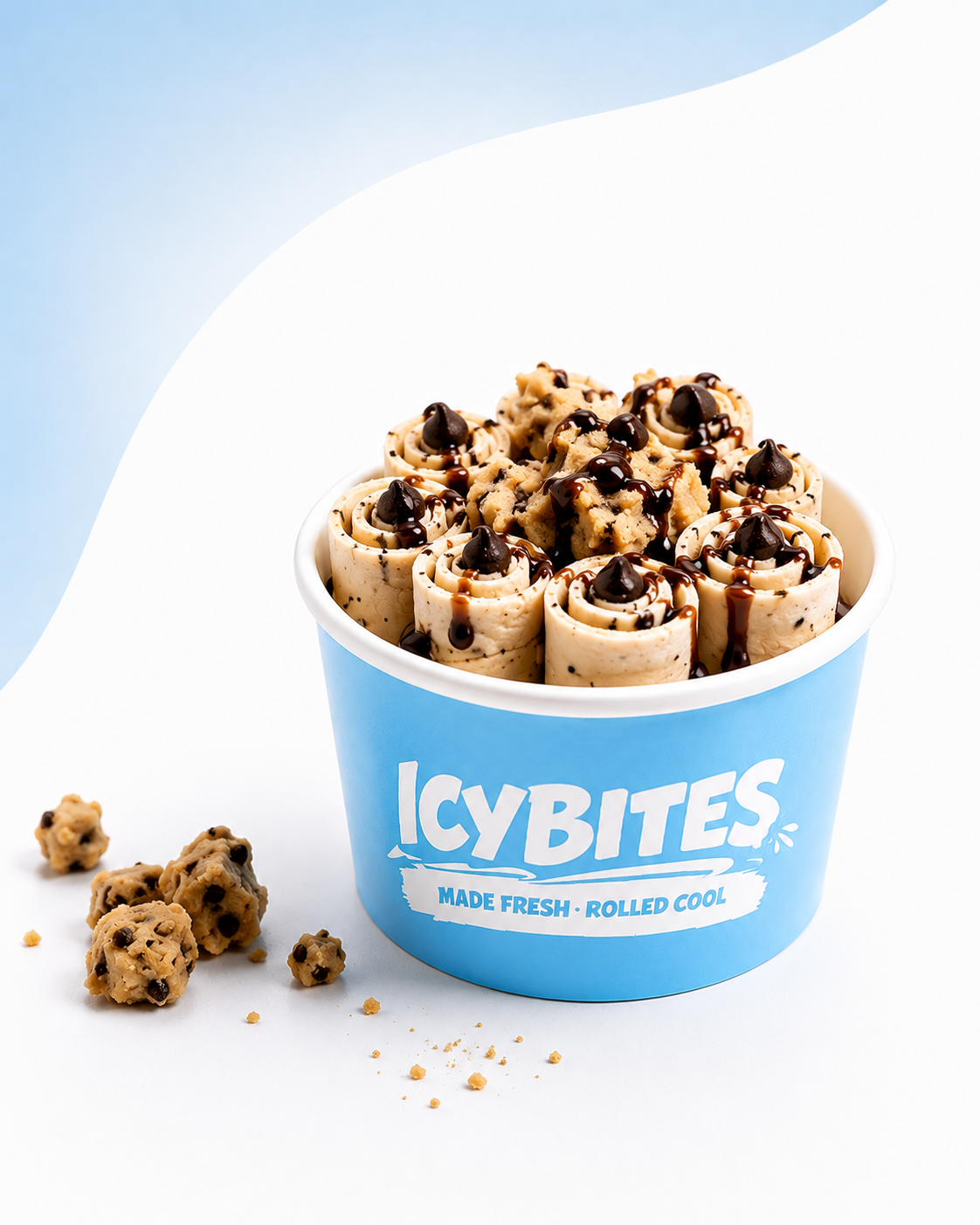 A blue ice cream cup filled with cookie dough ice cream rolled into spirals, topped with chocolate drizzle and cookie crumbs, with cookie dough pieces beside it on a white surface.