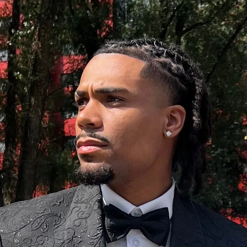 Partial profile of a man with braided hair, a diamond earring, dressed in a tuxedo with a bow tie, standing outdoors with trees and a red building in the background.