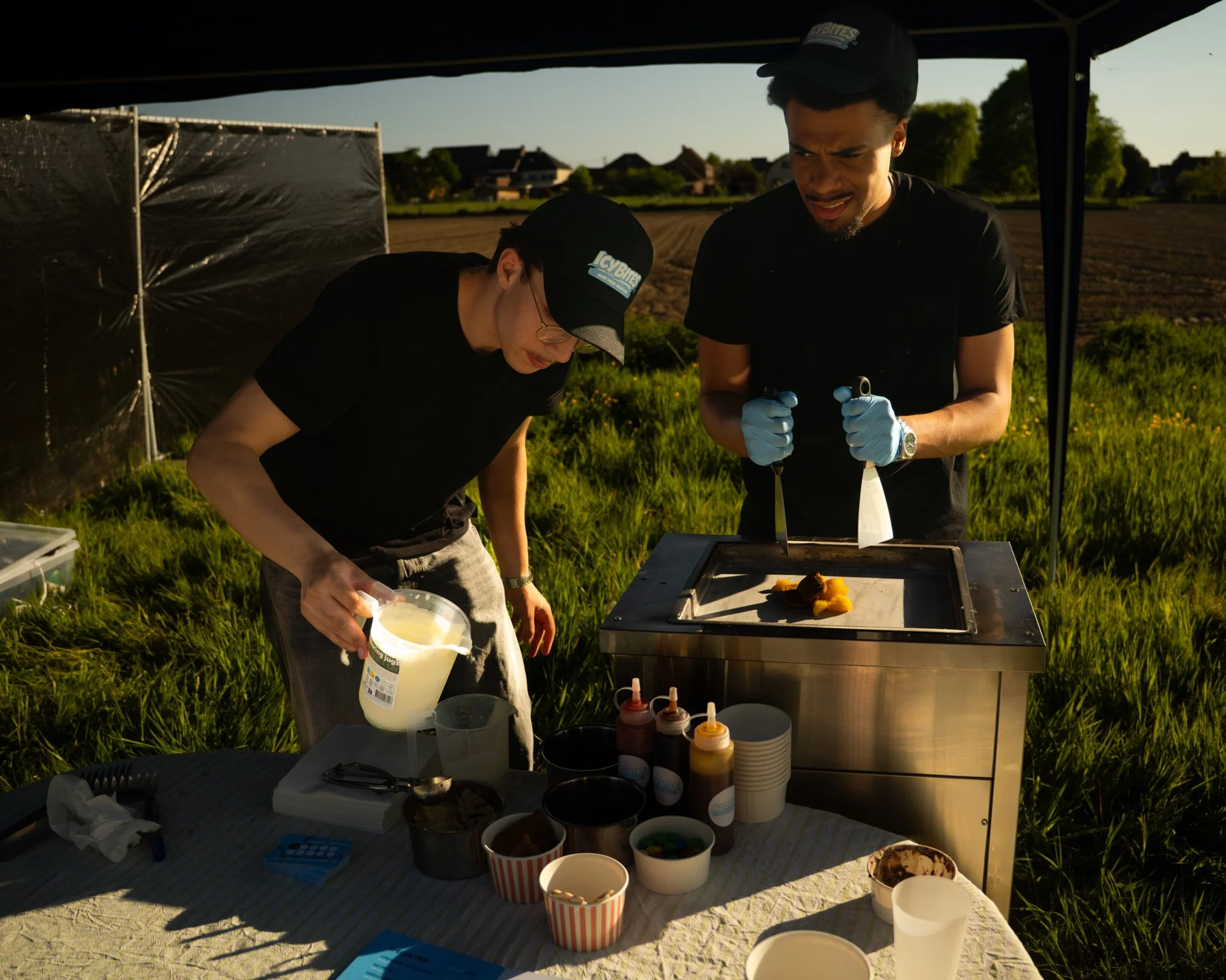 Two men working at an outdoor food stand, with various containers and toppings on a table, under a canopy on a grassy area during sunset.