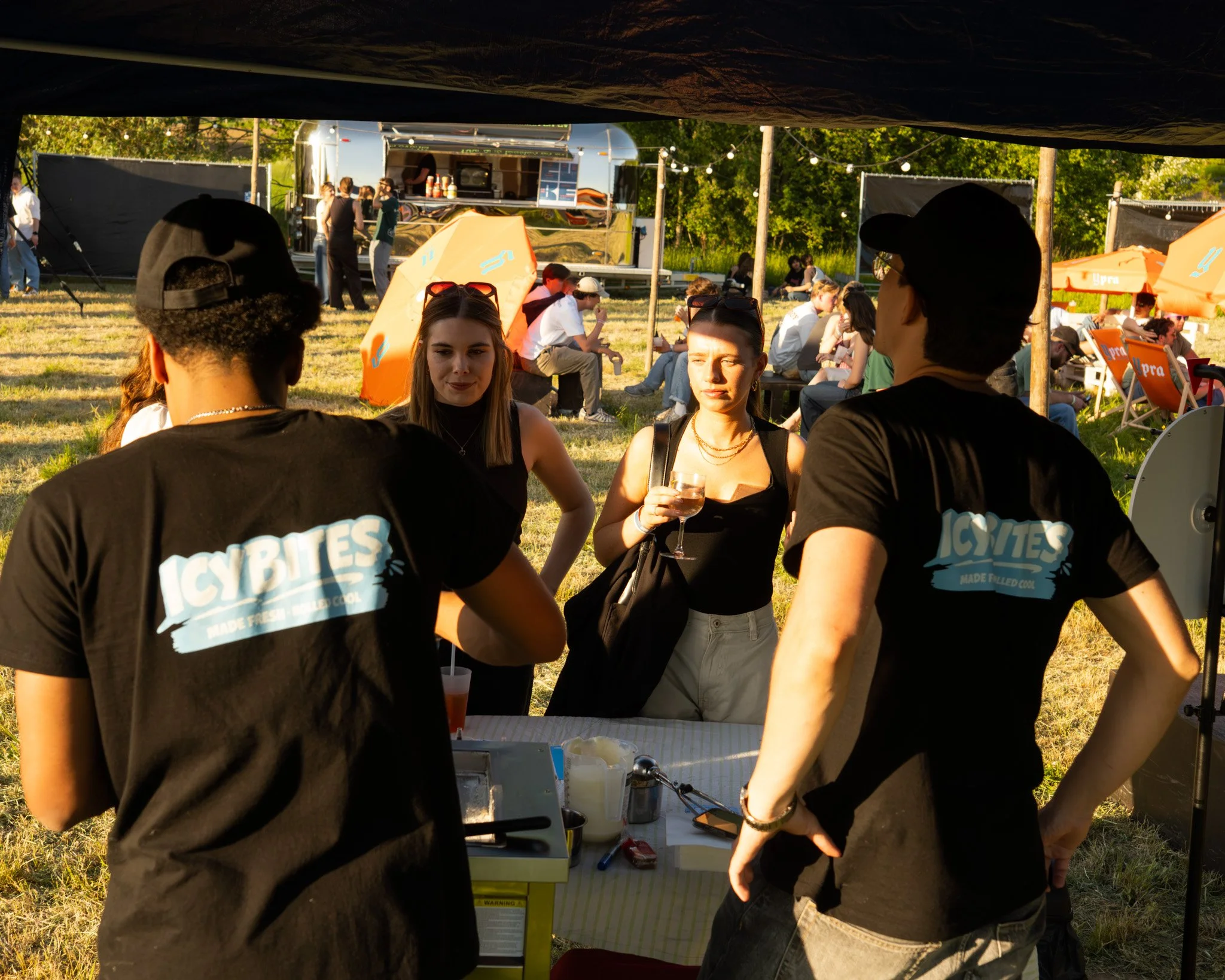 People gathered at an outdoor event seen through a tent opening, with a food truck, string lights, and trees in the background.