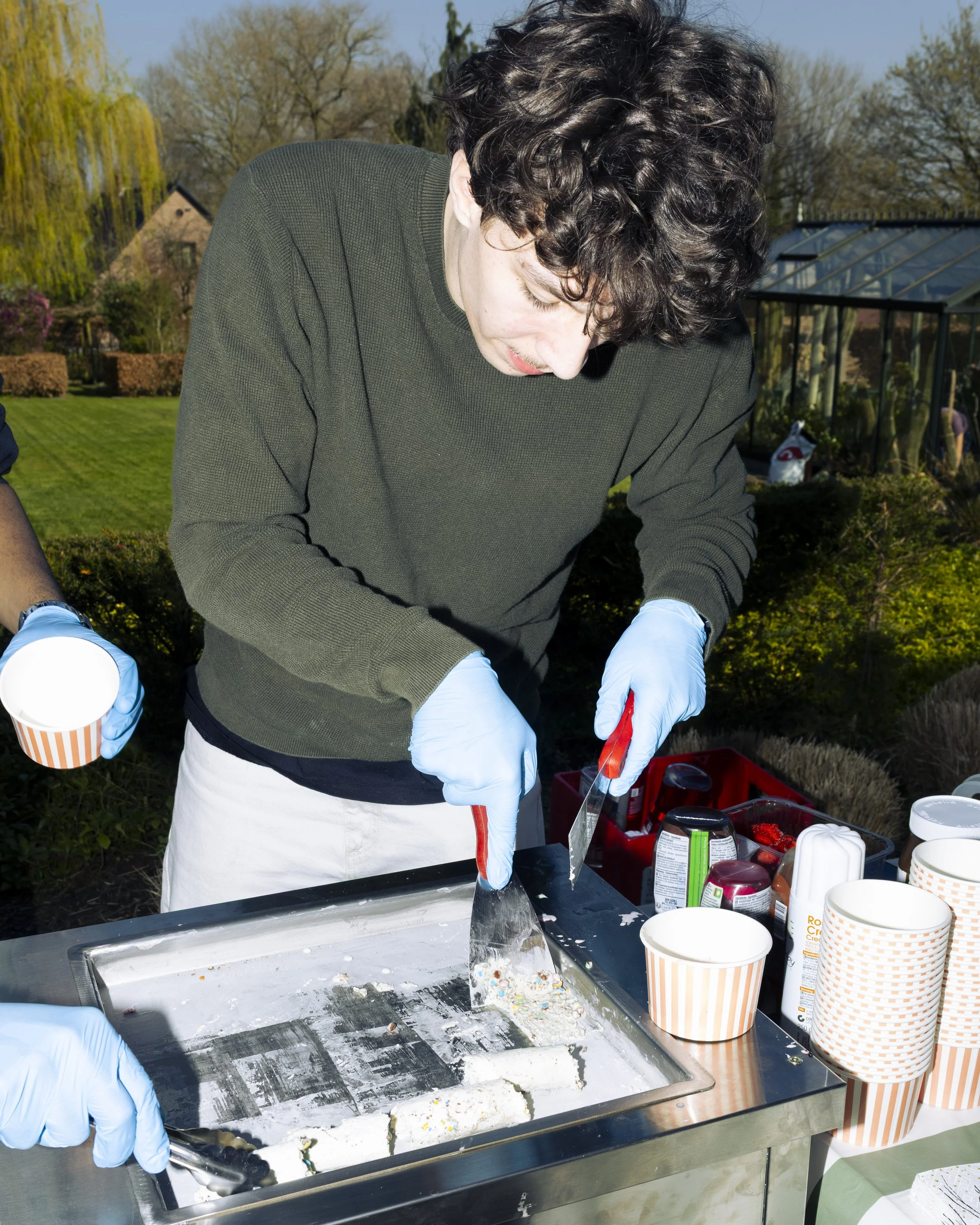 A person wearing a dark green sweater and white gloves appears to be preparing or serving ice cream or frozen desserts at an outdoor ice cream stand. There are empty striped paper cups, containers of toppings, and an ice cream machine on the stainless steel counter. In the background, there are trees, a greenhouse, and a garden under a clear blue sky.