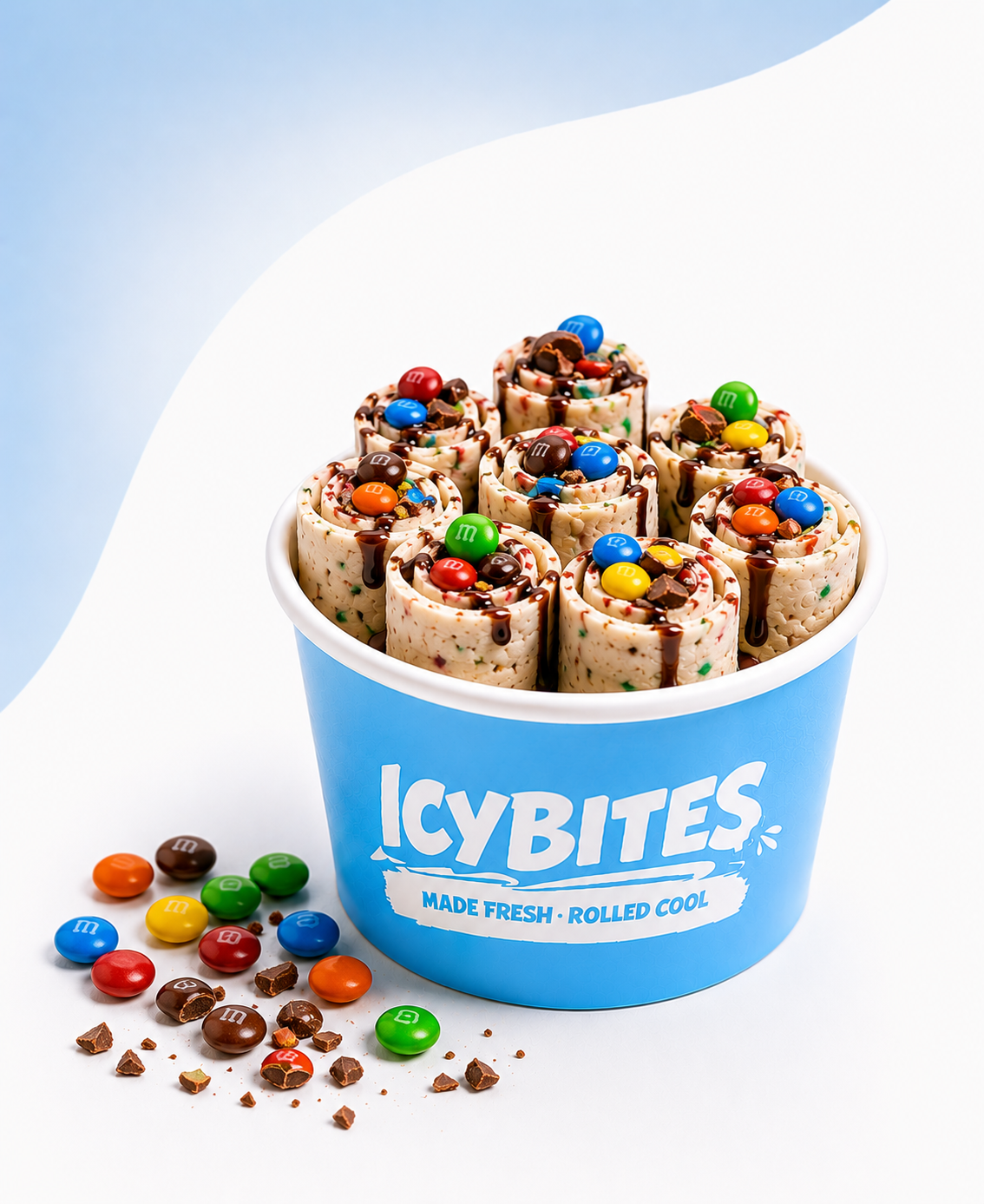 A blue bucket labeled 'ICY BITES' filled with rolled ice cream swirls topped with M&M candies and chocolate drizzle, with some candies and chocolate pieces spilled on the white surface nearby.