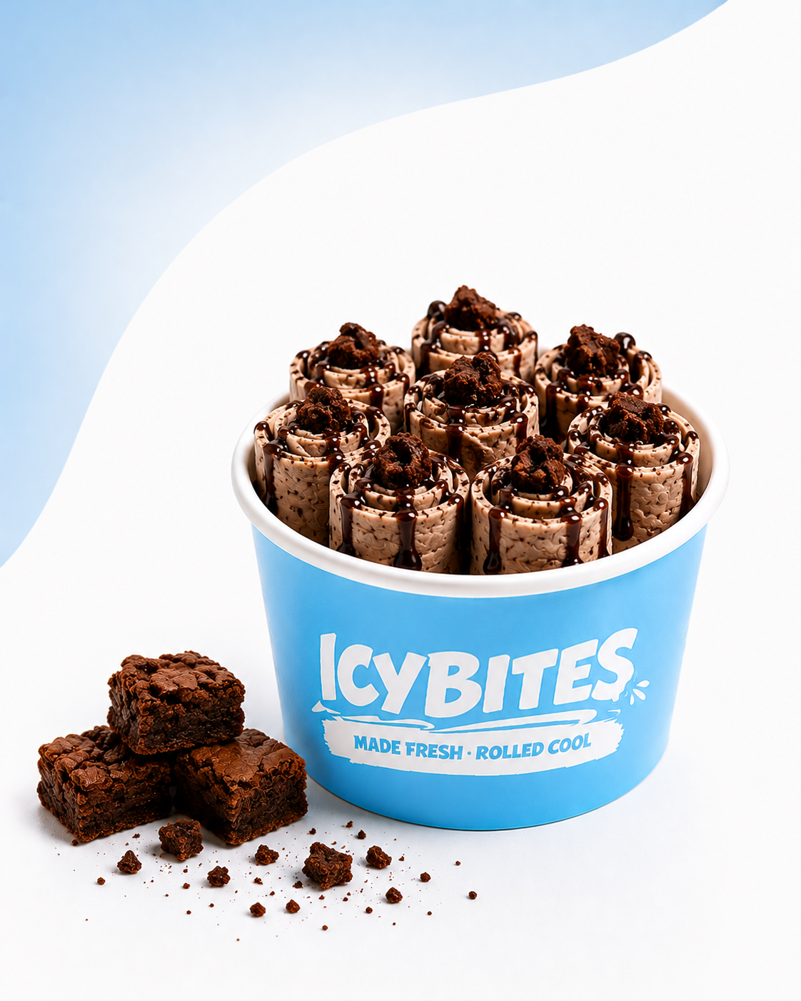 A blue bucket labeled "ICYBITES" filled with chocolate ice cream rolls topped with chocolate syrup and cookie crumbles, with chocolate brownie pieces and crumbs beside the bucket.