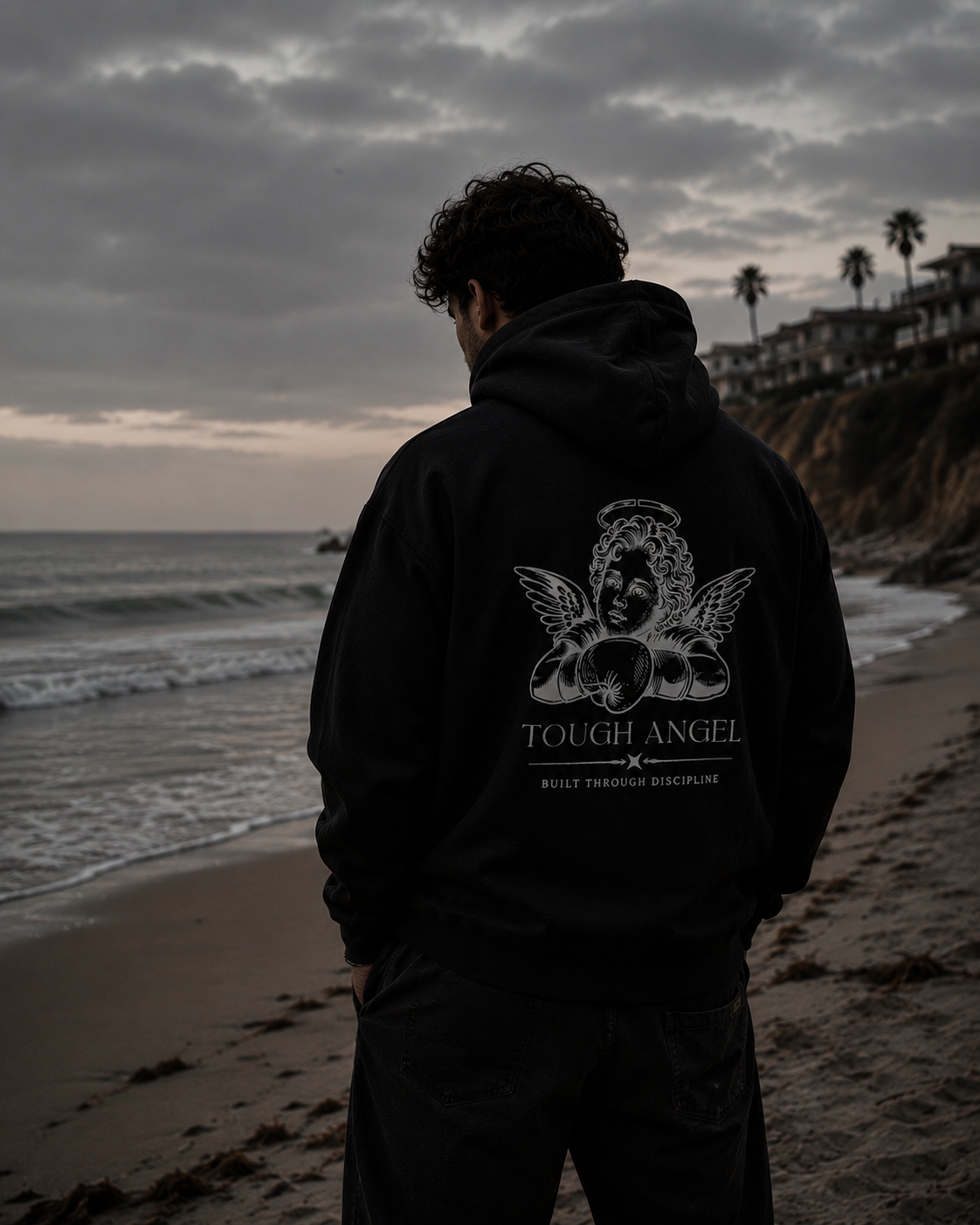 A man standing on a beach facing the ocean with his back to the camera, wearing a black hoodie with a graphic of a winged angel and the words 'Tough Angel' and 'Built Through Discipline.' The sky is cloudy and there are houses on a hill with palm trees in the background.
