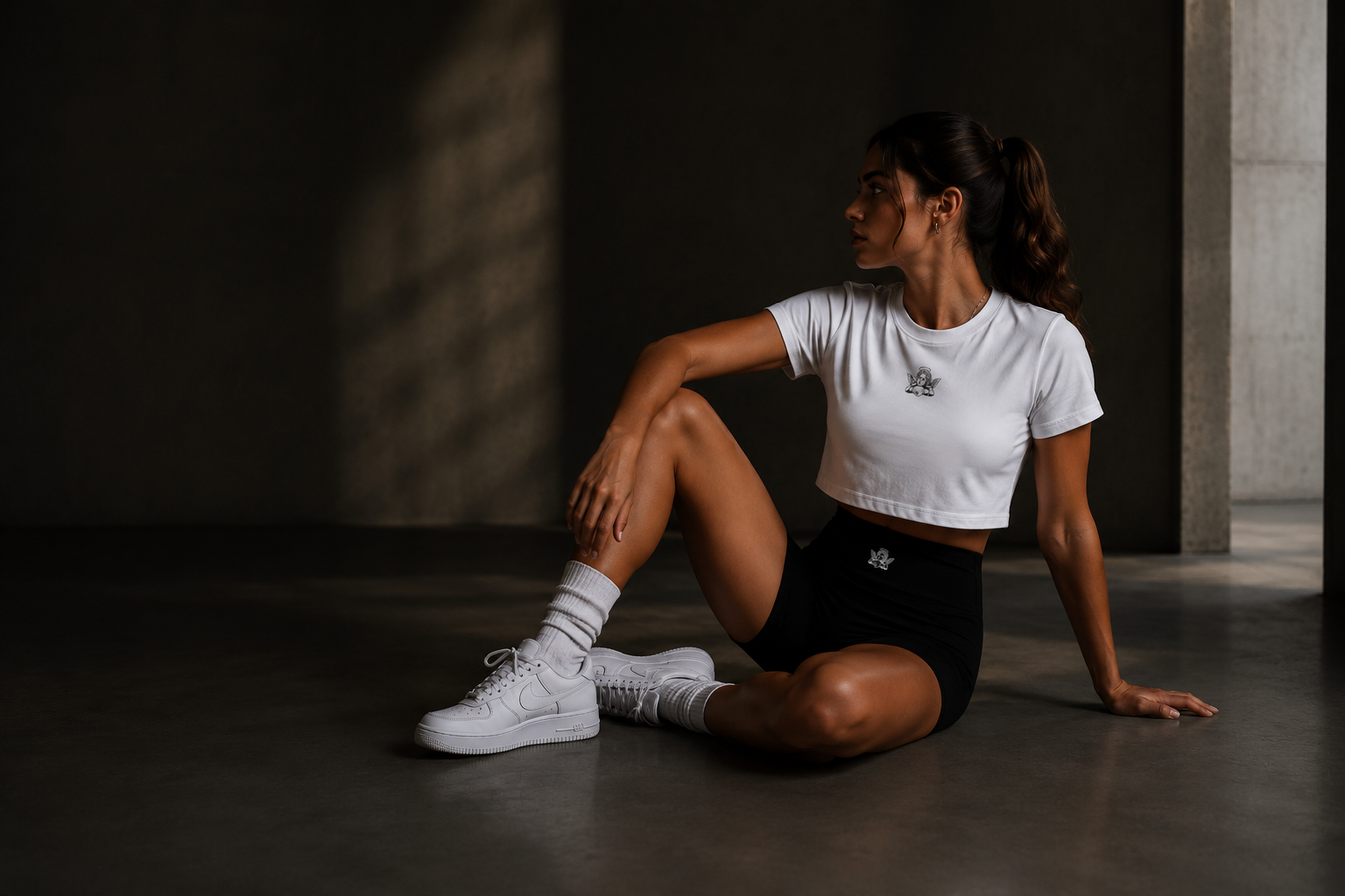 A woman in athletic clothing sitting on the floor against a dark wall, looking to the side with her hand on her knee.