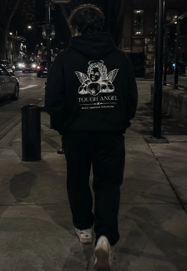 Person walking on a city sidewalk at night, wearing a black hoodie with a design of a winged angel and boxing gloves on the back, urban street with parked cars and streetlights.