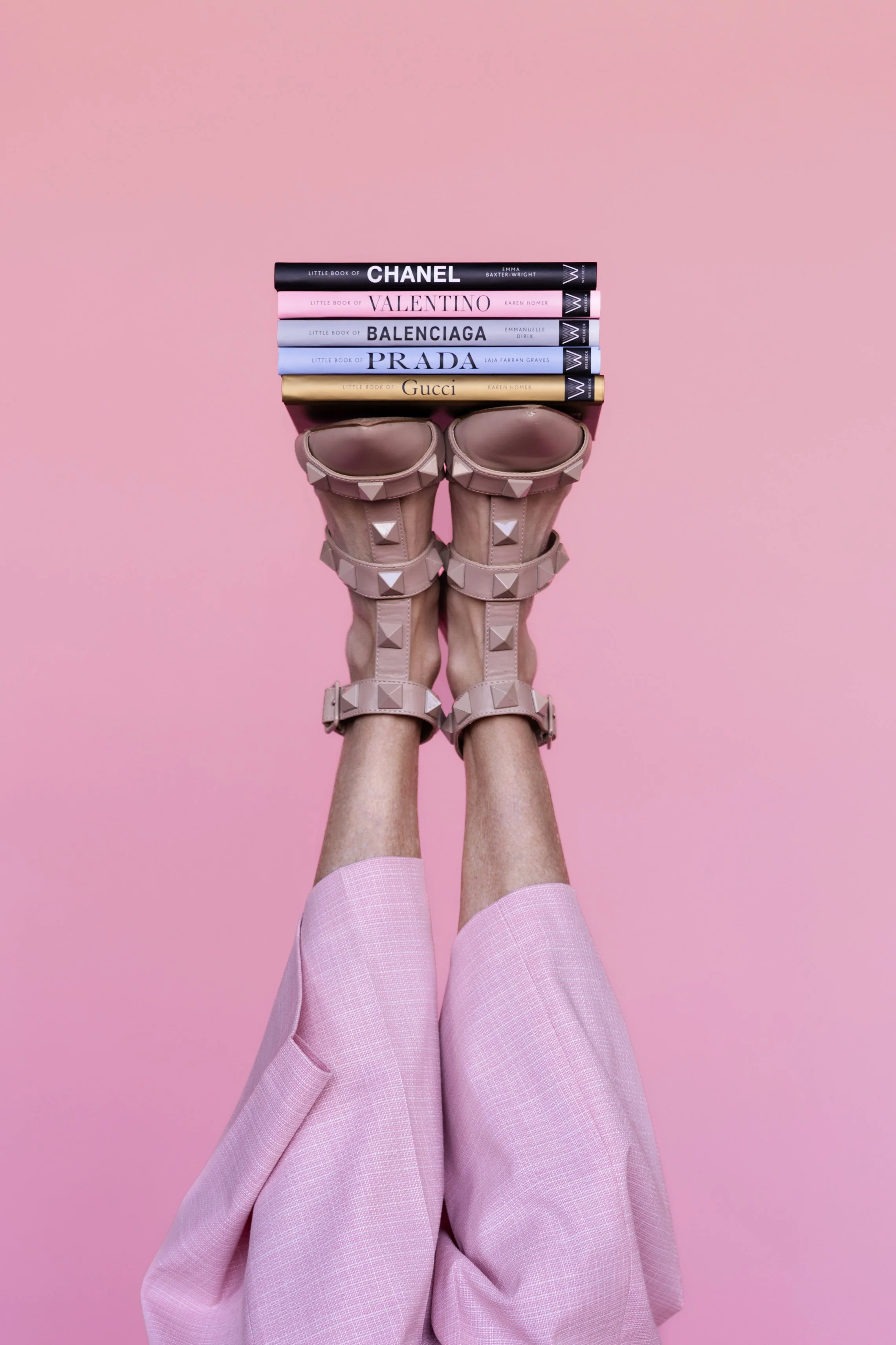 Person holding a stack of fashion books while wearing light pink skirt and nude high-heeled shoes with studded straps on a pink background.