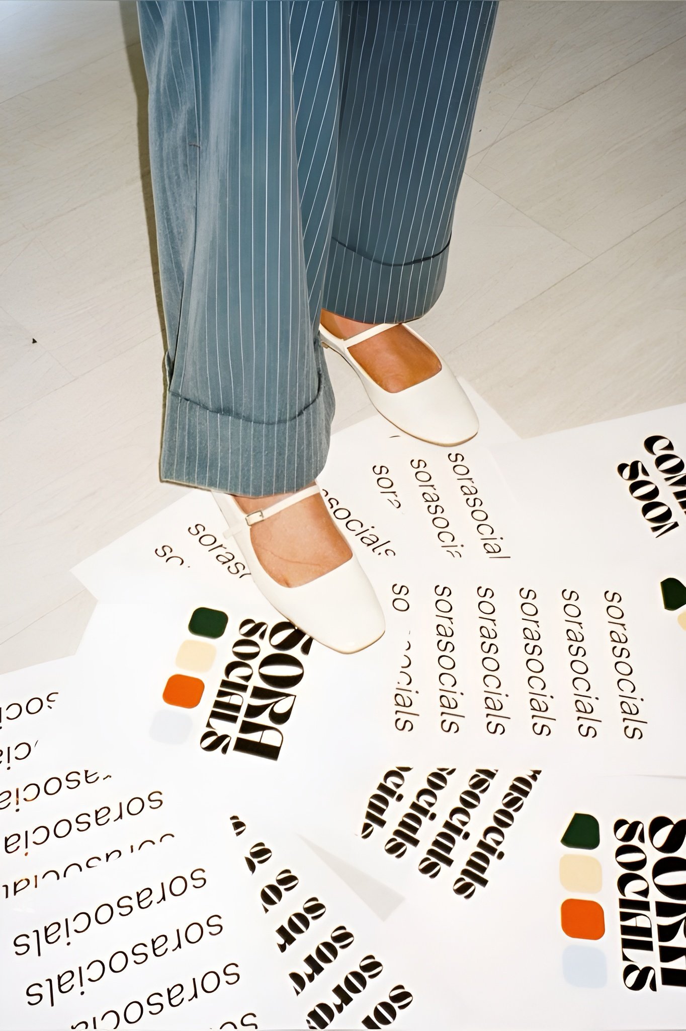 Close-up of a mannequin's legs in gray pinstripe pants and white shoes standing on a sheet of paper with the word 'socials' repeated, with a color palette featuring green, beige, orange, and black.