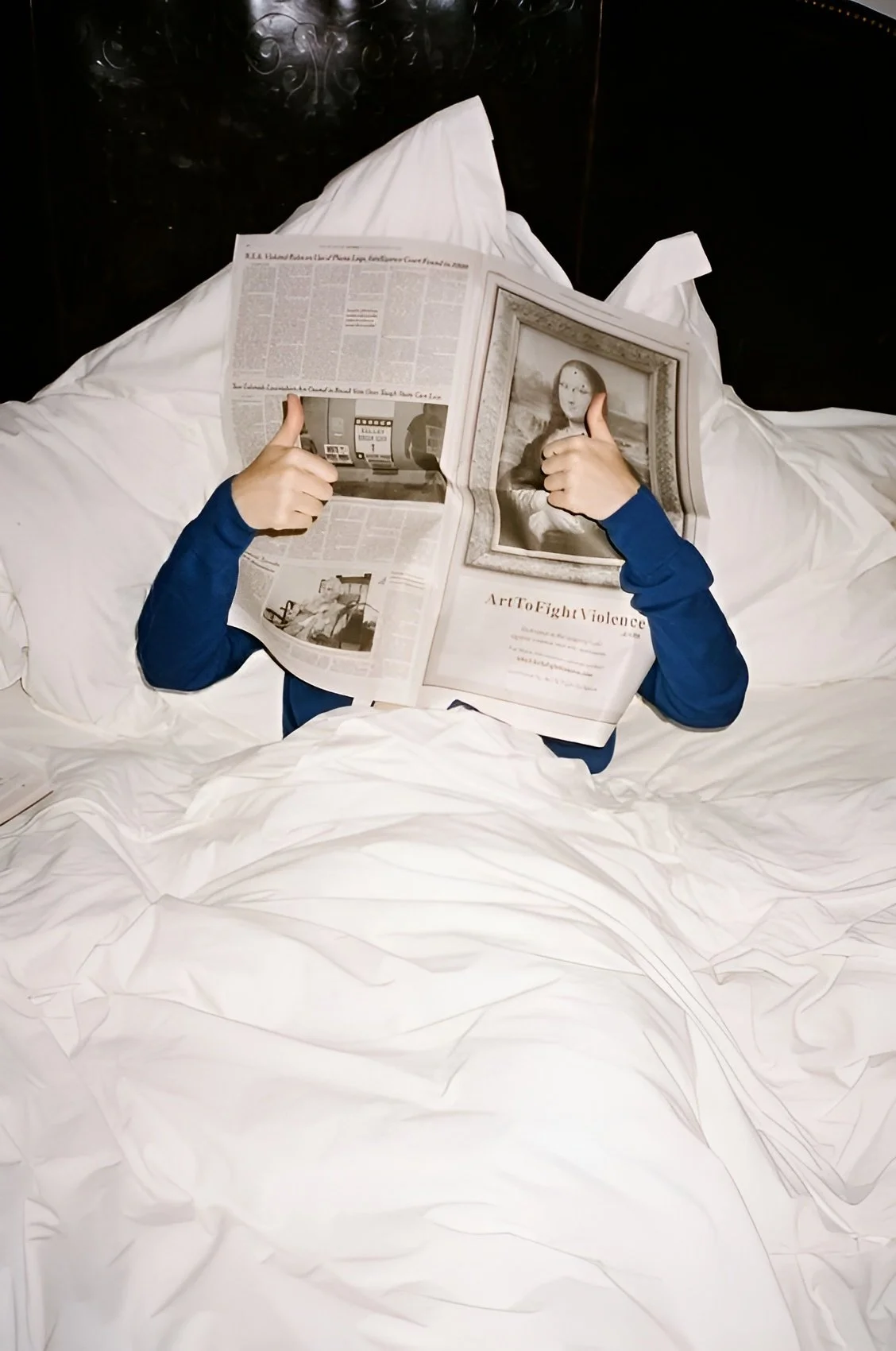 Person lying in bed reading a newspaper with a framed art piece titled 'Art To Fight Violence' on it.