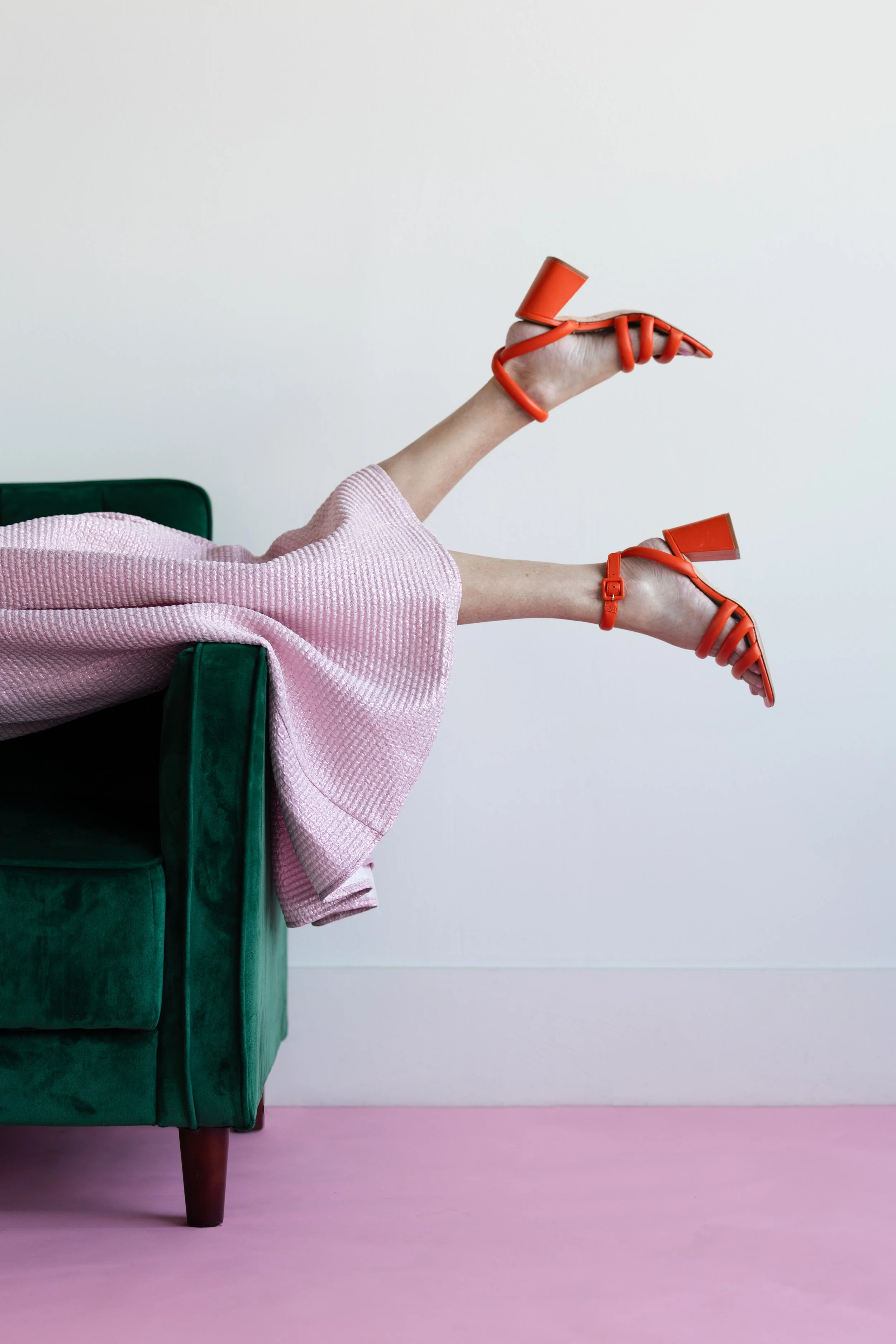 Person lying on a green velvet couch with legs raised in the air, wearing orange high heels and a pink textured outfit, against a plain white wall and pink floor.