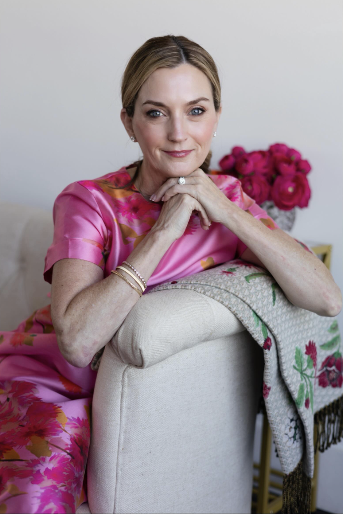 A woman with blonde hair and blue eyes sitting on a beige sofa, wearing a pink floral dress, with pink flowers in a vase behind her.