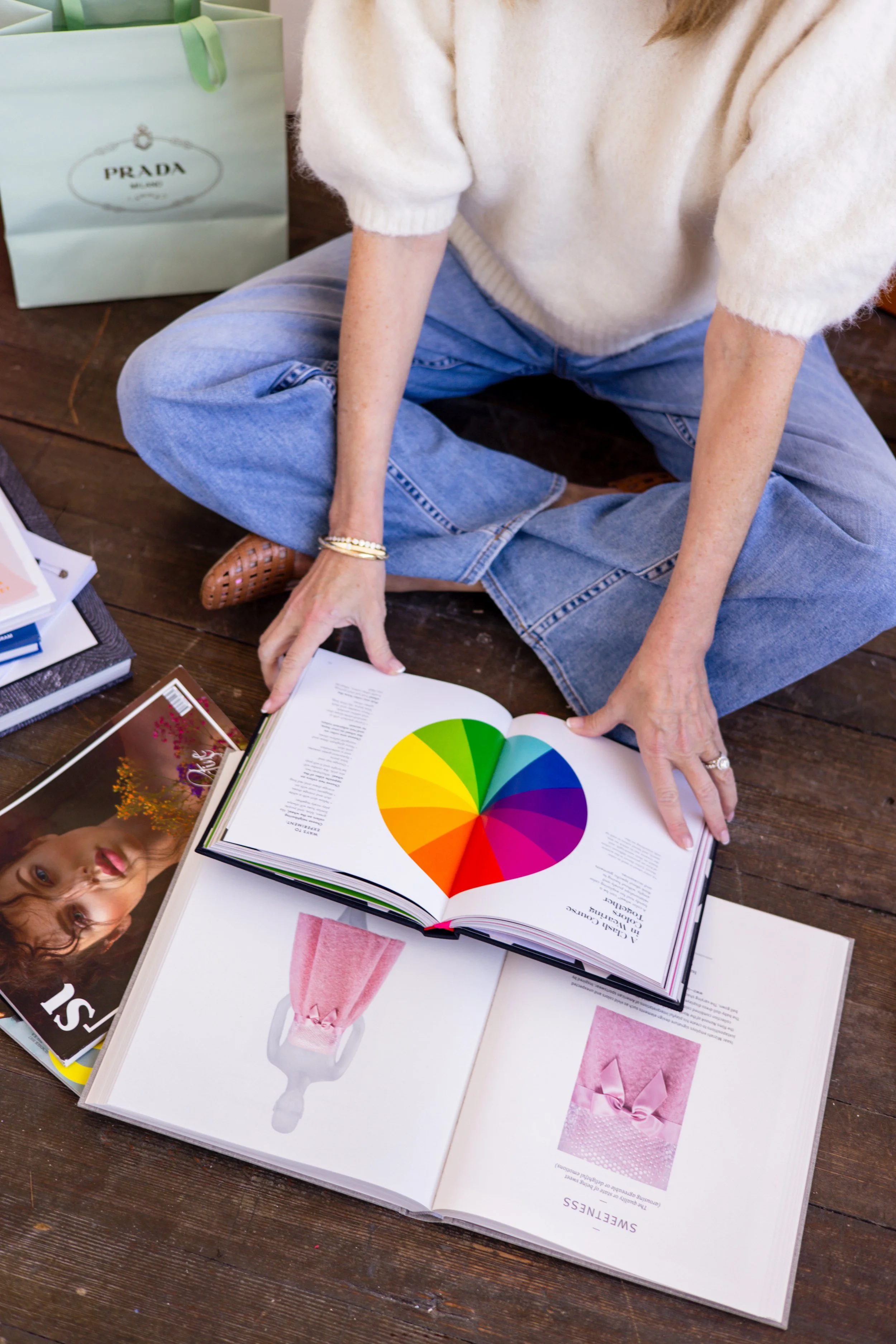 Close-up of a person's lap as they sit on the floor, with open magazines and books, including a color wheel and images of gifts, a pink drink, and a woman, on a wooden surface.