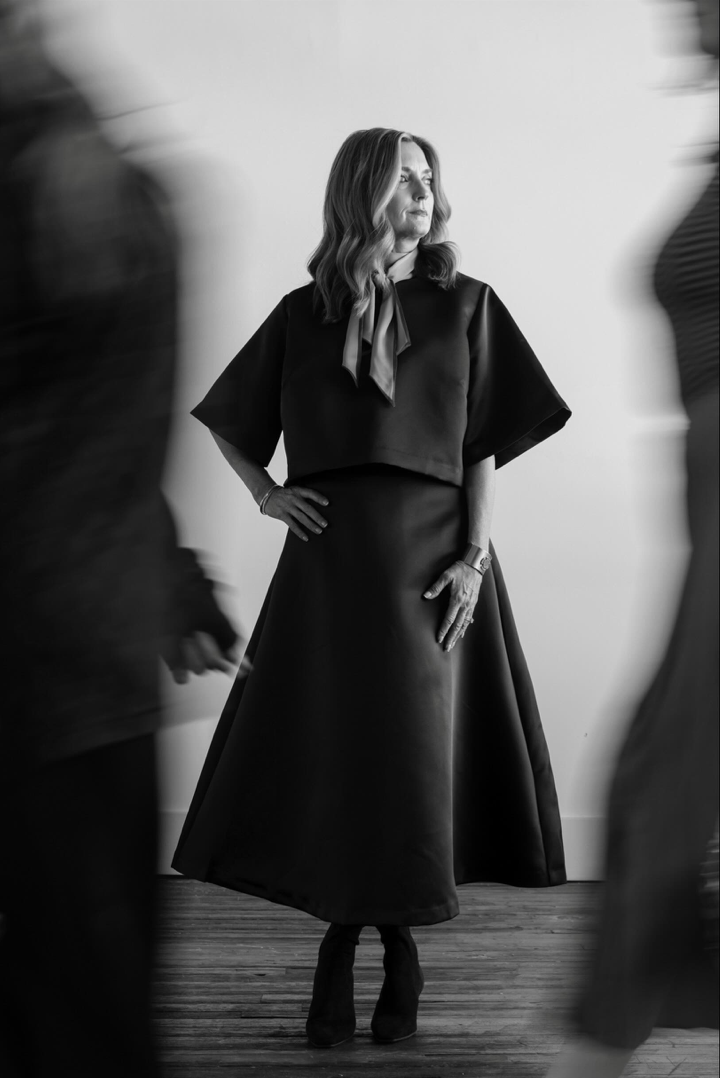A woman with shoulder-length hair wearing a dark dress with a layered skirt and a silk scarf is standing with her hand on her hip, looking to her left. Two blurred figures are walking past in the foreground.