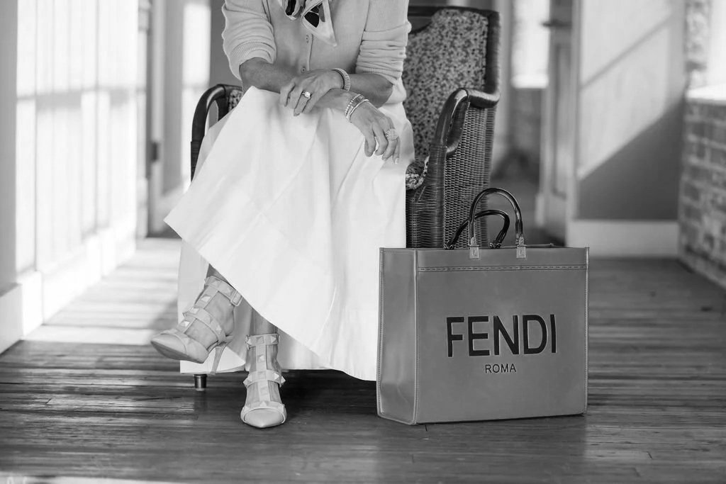 A woman sitting on a chair, wearing a white dress and high boots, with a Fendi shopping bag placed on the floor beside her.
