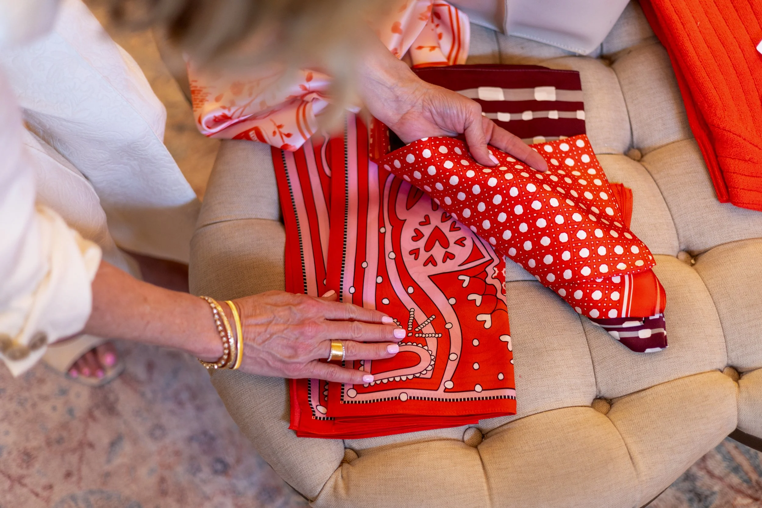 Close-up of a person sewing red and white patterned fabric, with another person nearby helping or observing, on a beige tufted cushion.