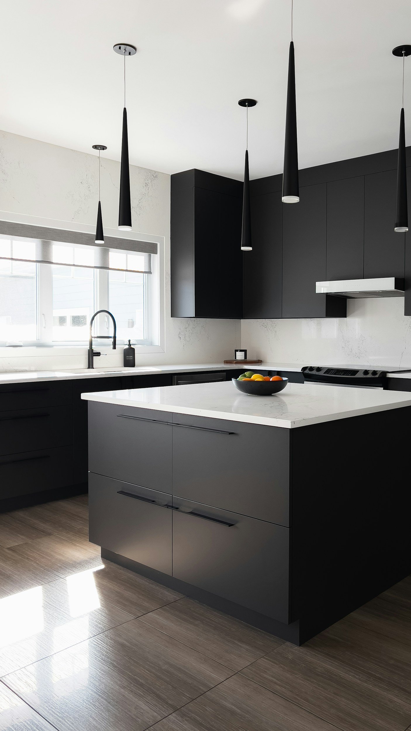 Modern kitchen with black cabinets, a white marble island, a bowl of fruit, large window, and pendant lights.
