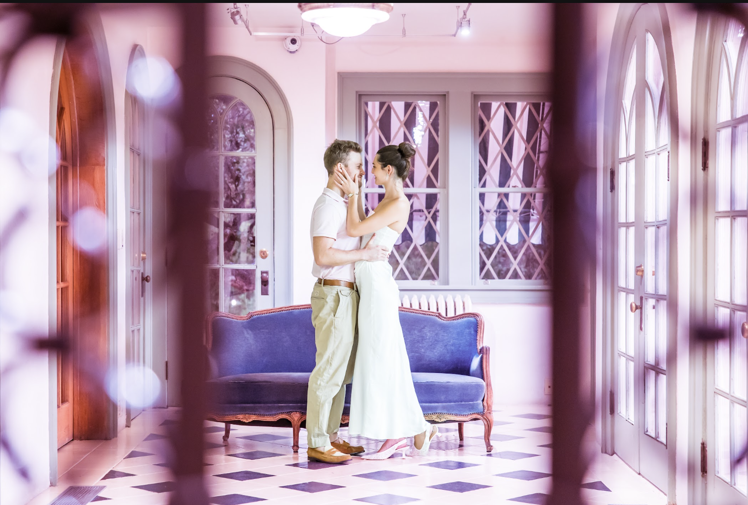 A couple in wedding attire standing close indoors, touching foreheads, framed by a decorative gate or railing, with a pink-toned elegant room featuring large windows and a vintage blue sofa in the background.