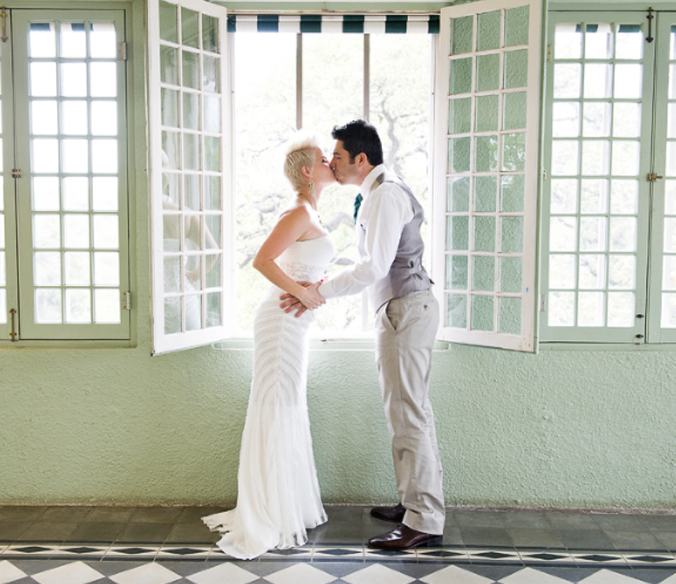 A couple sharing a kiss in front of open windows with green frames, bright natural light, and a tiled black and white floor.