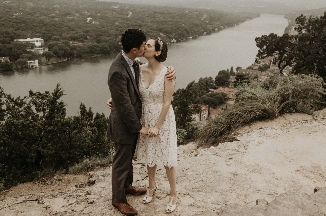 A couple in wedding attire sharing a kiss on a hillside overlooking a river and lush landscape.