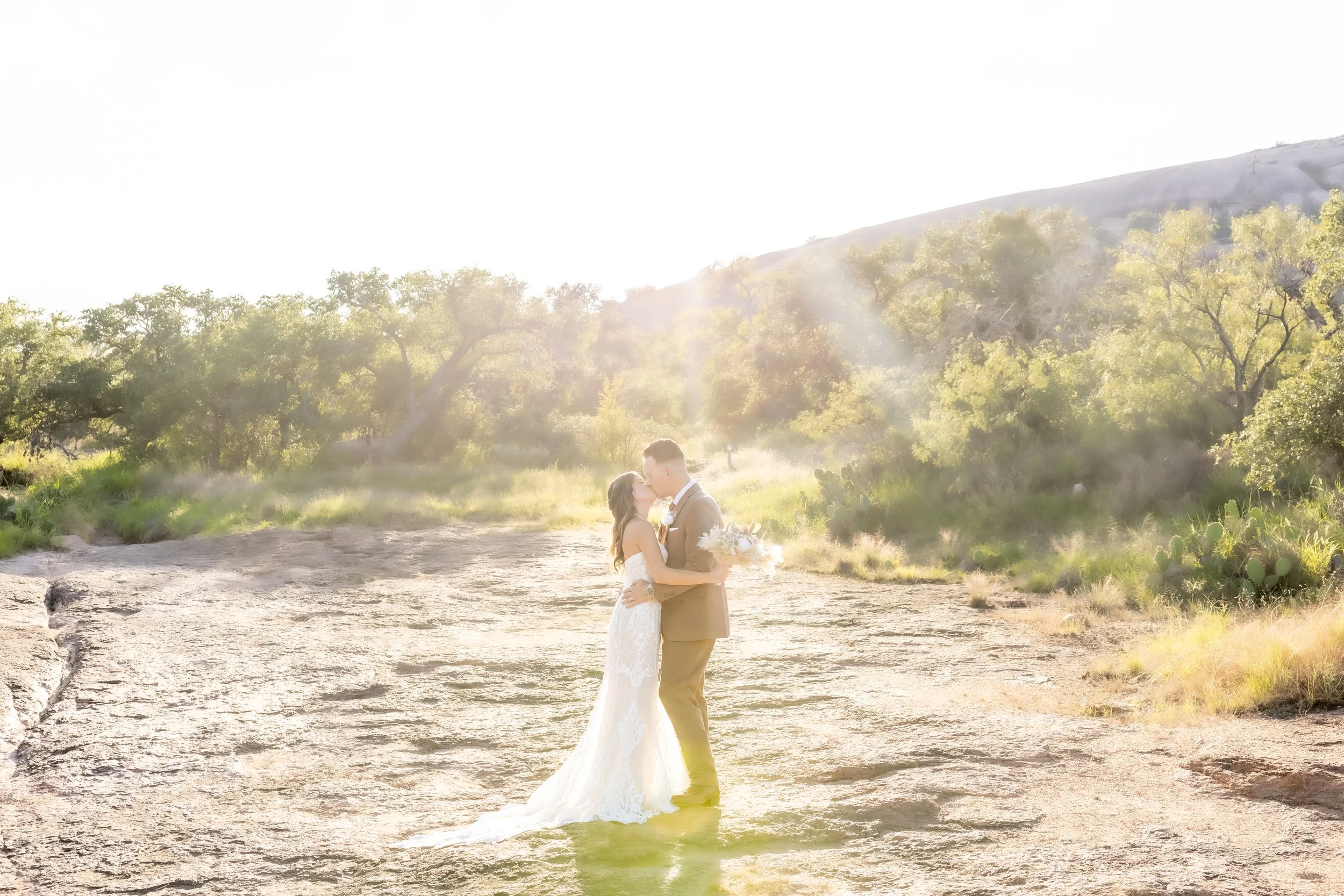 A bride and groom sharing a kiss outdoors during sunset, with trees and hills in the background.