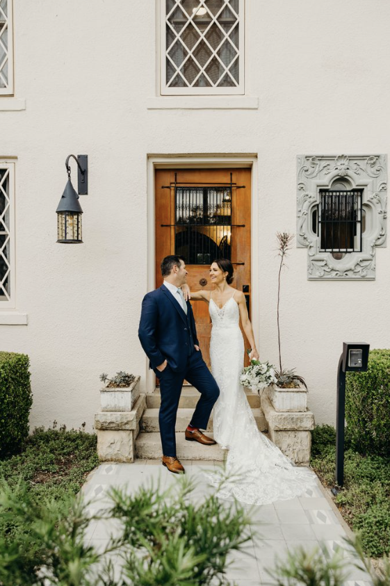 A bride and groom standing in front of a house door on their wedding day. The bride is wearing a white wedding dress and holding a bouquet, while the groom is dressed in a navy blue suit. They are smiling and looking at each other.
