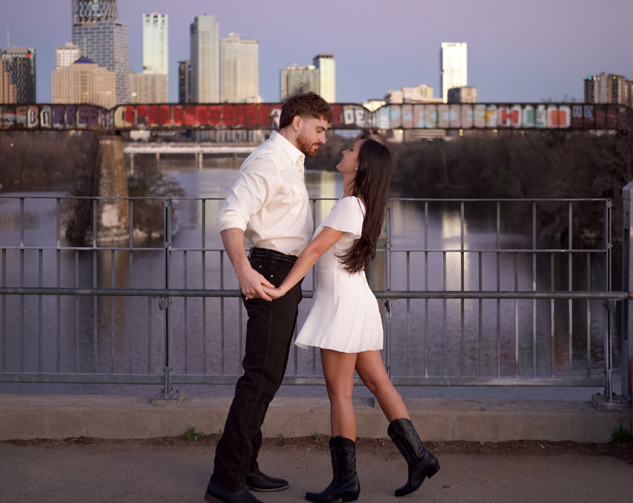 A young couple holding hands faces each other on a bridge with city skyline and graffiti-covered bridge in the background during sunset.