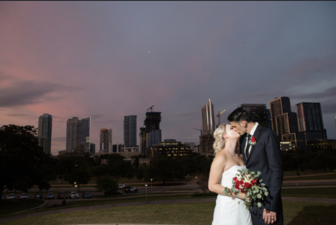 A bride and groom kissing outside during sunset with a city skyline in the background.