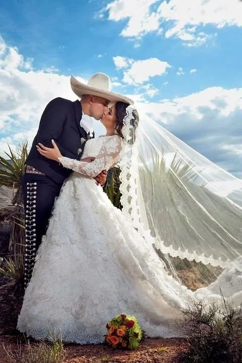 A couple dressed in traditional Mexican attire sharing a kiss outdoors on a sunny day with a partly cloudy sky.