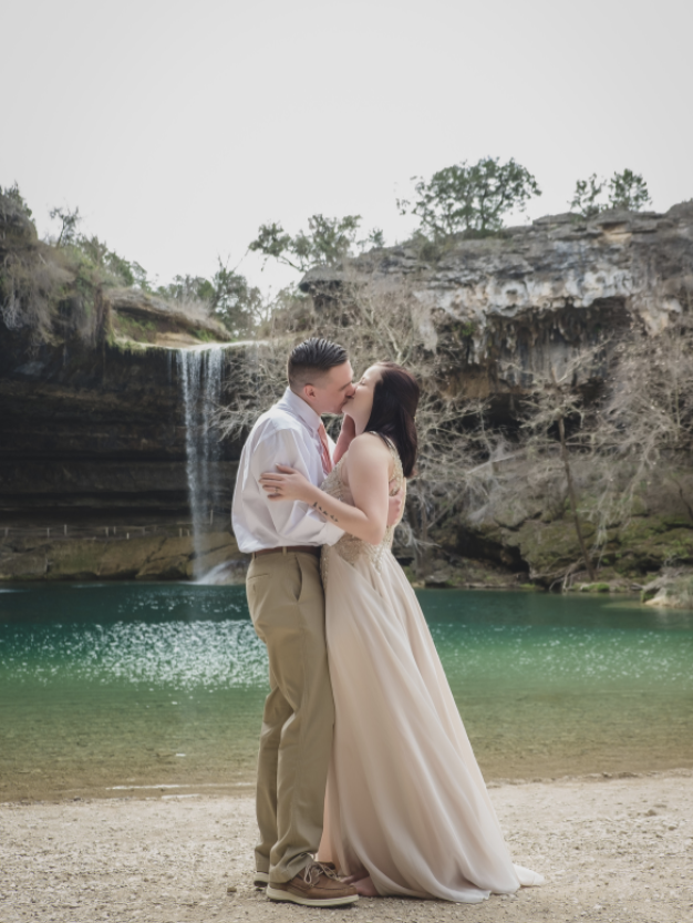 A couple kissing on a beach near a waterfall and rocky cliffs, with trees in the background.